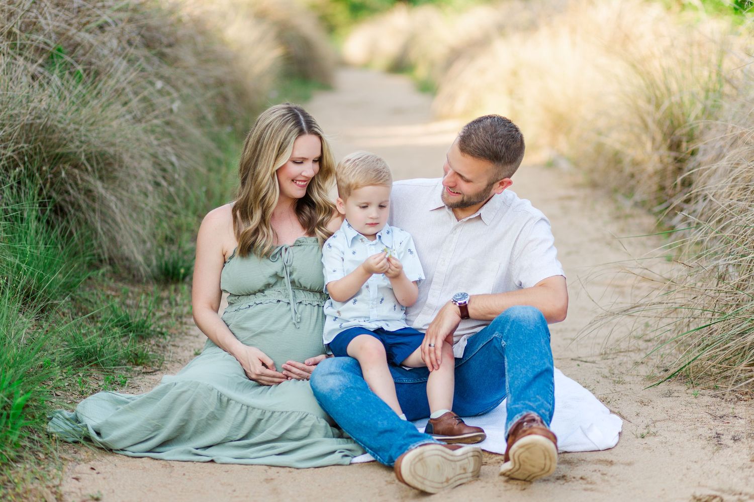 A family sits together on a dirt path surrounded by tall grass during a summer outdoor photo session.