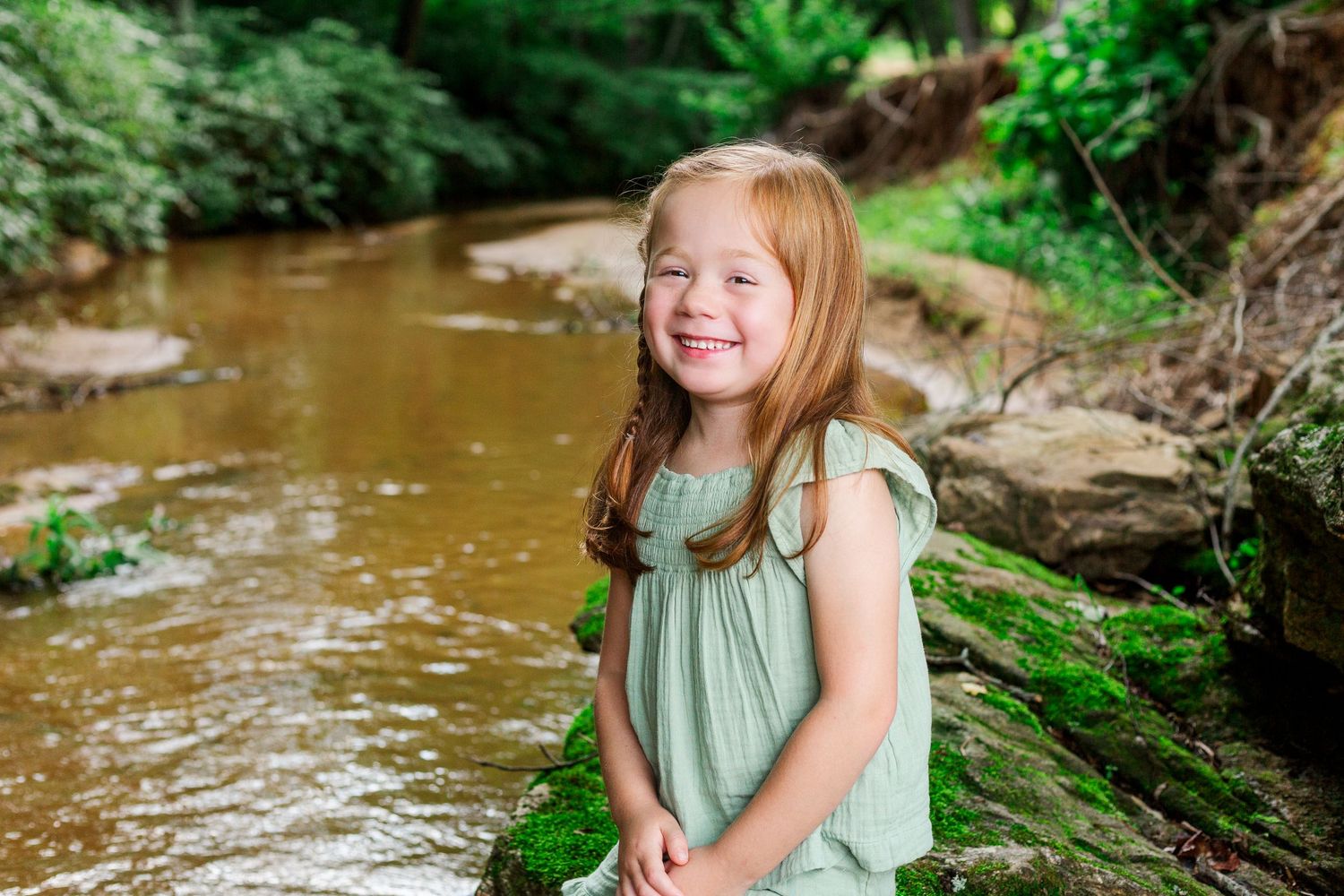 Young child in a mint green outfit smiles while playing in a shallow forest stream.