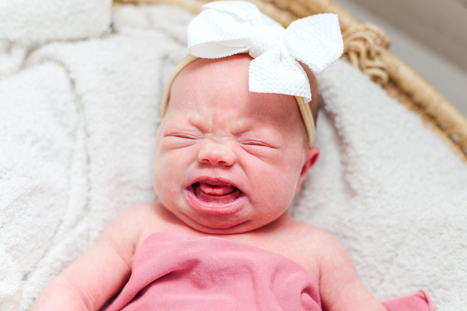 Crying newborn baby wearing white bow headband lying on soft beige blanket.