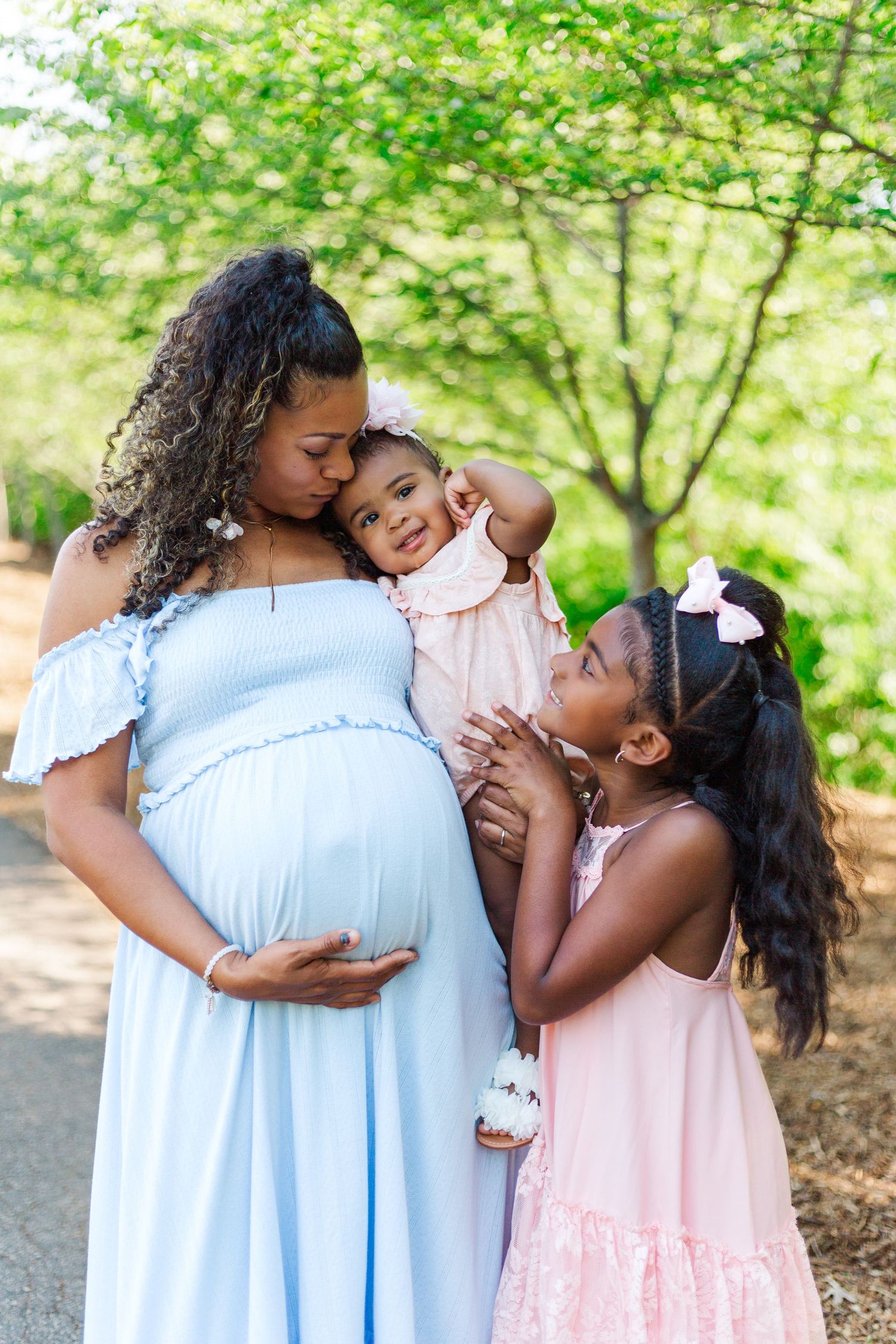 Pregnant woman in blue dress kisses toddler while older daughter in pink dress touches her baby bump outdoors.