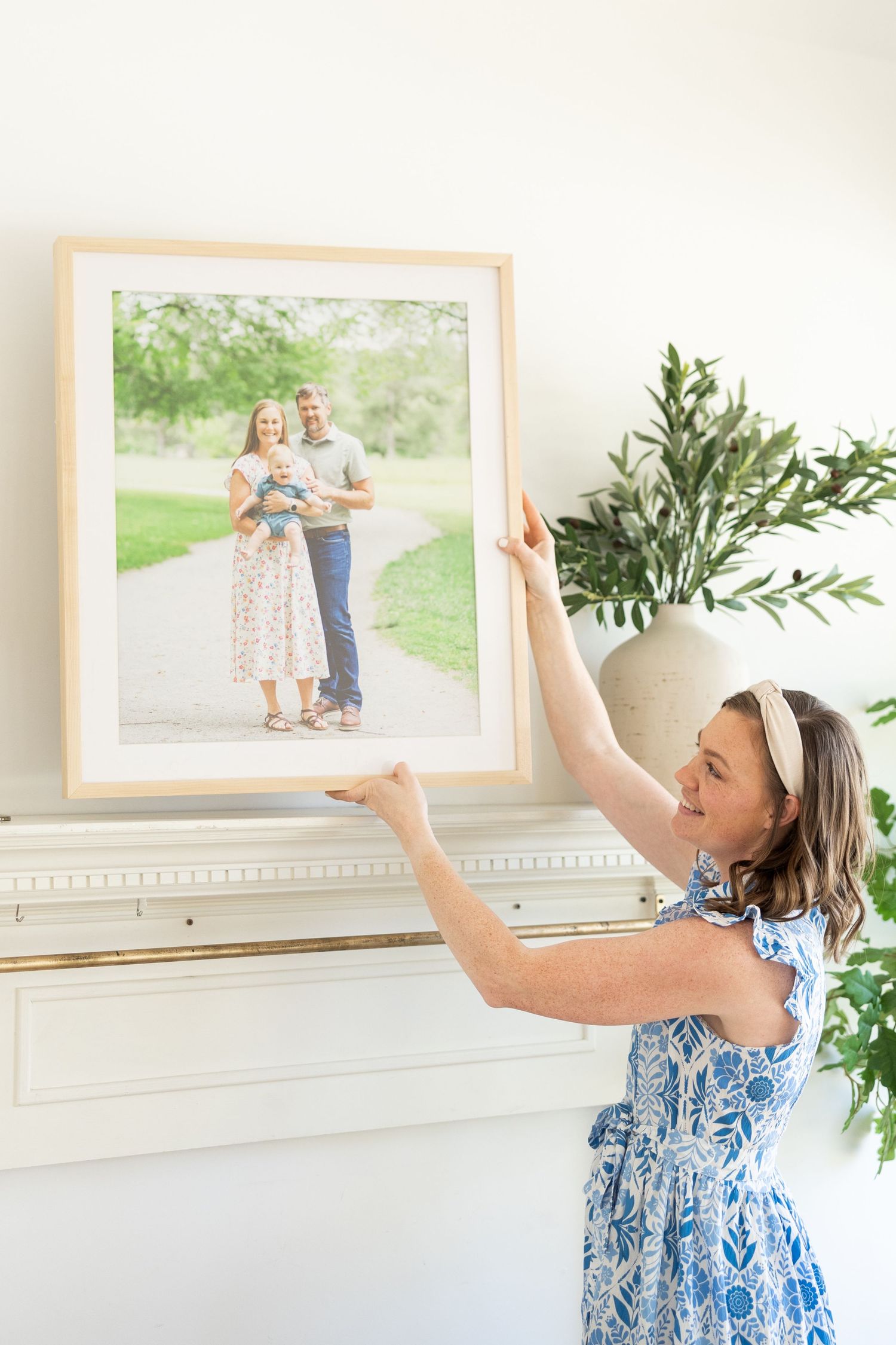 Person in blue and white patterned dress hanging framed photograph on white wall next to decorative plant.