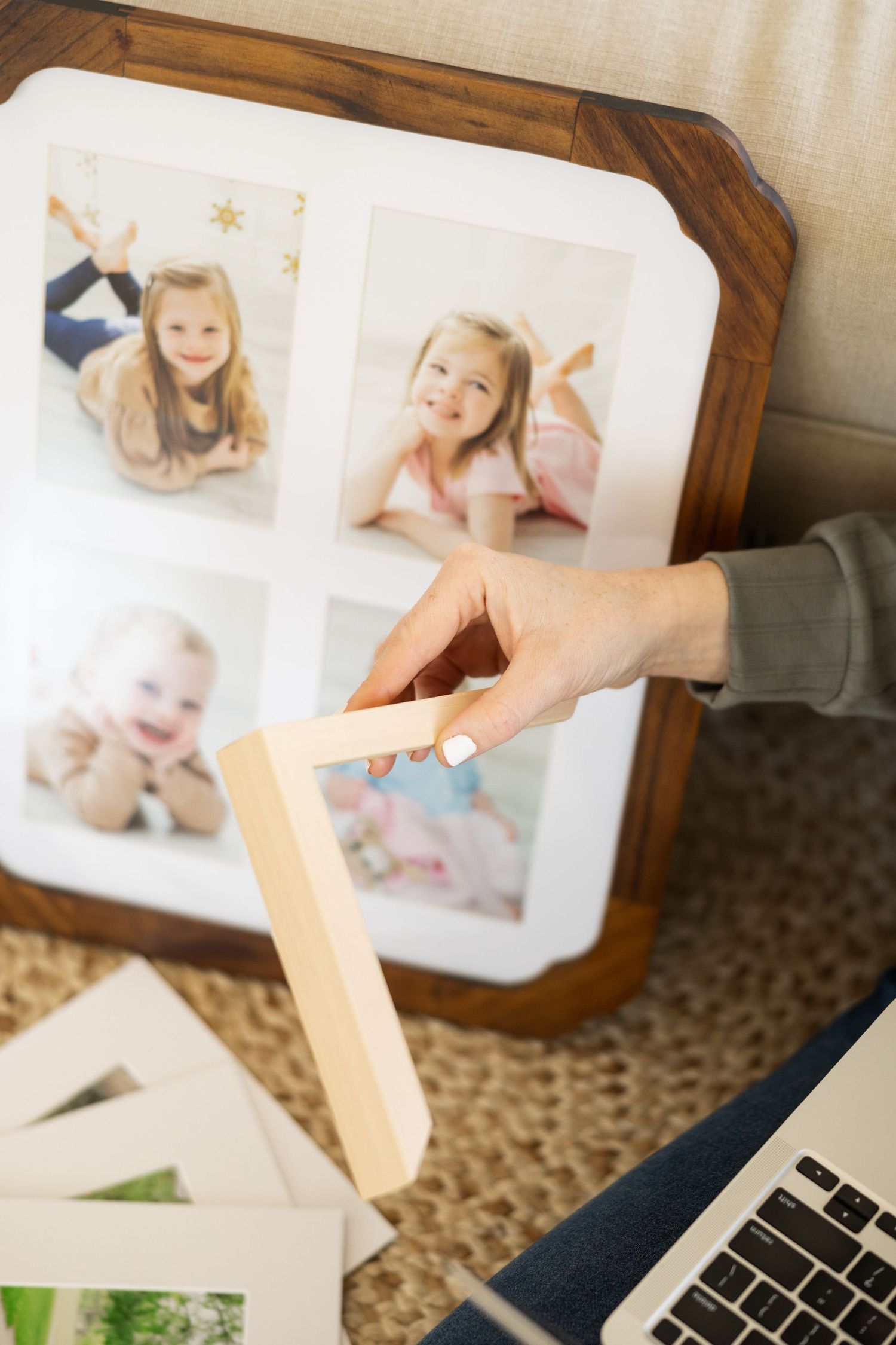 A hand holds a wooden frame while looking at photographs displayed in a brown frame next to a laptop on a carpeted floor.