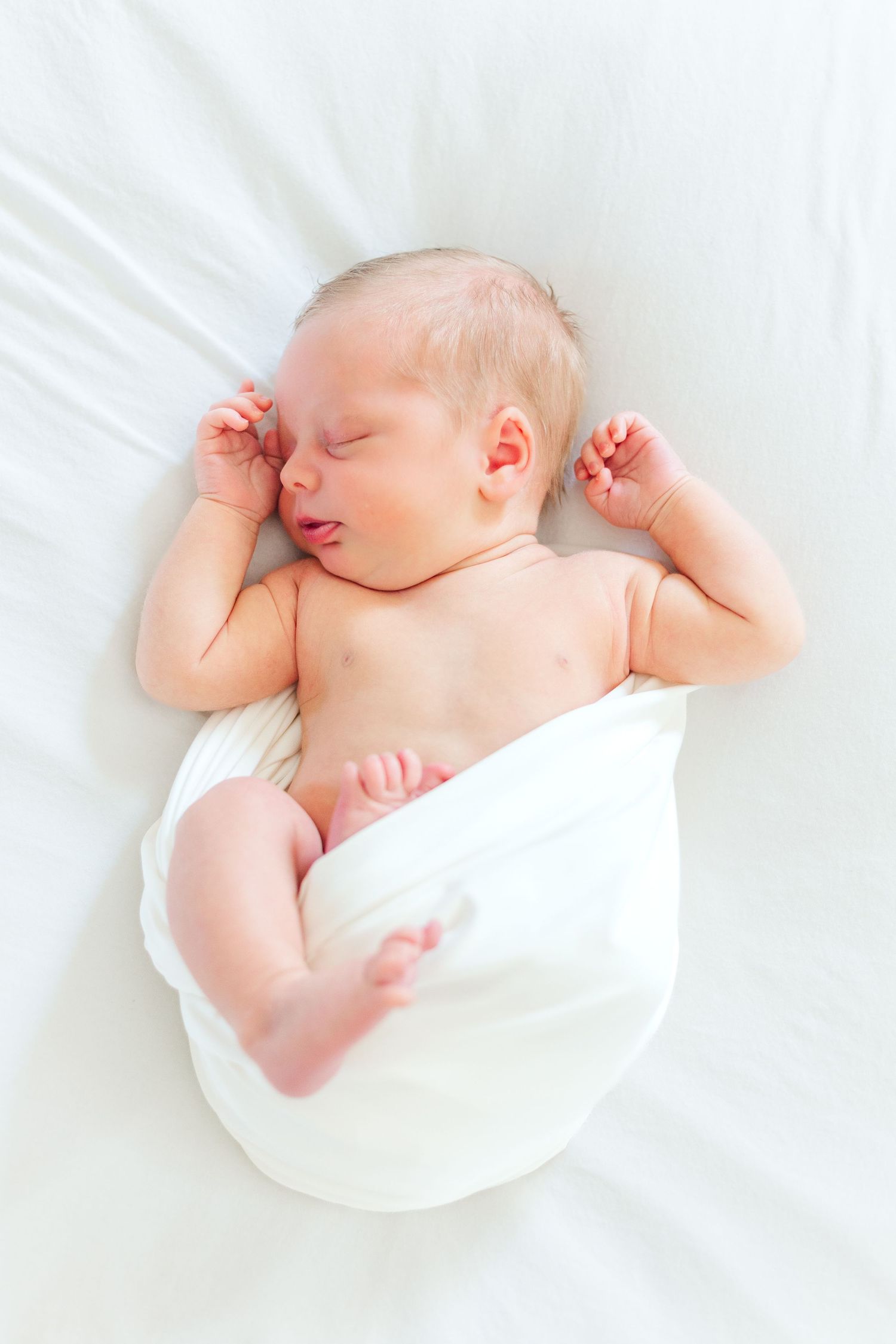 A peaceful newborn baby sleeping on white bedding, partially wrapped in a soft white blanket with tiny hands raised.