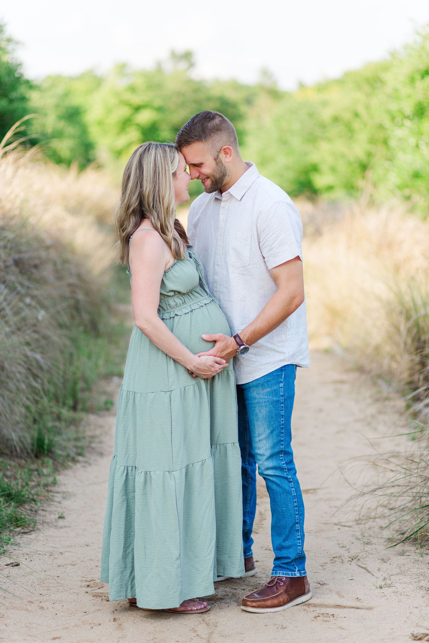 A couple shares an intimate moment on a dirt path surrounded by tall grass and greenery as they await their new arrival.