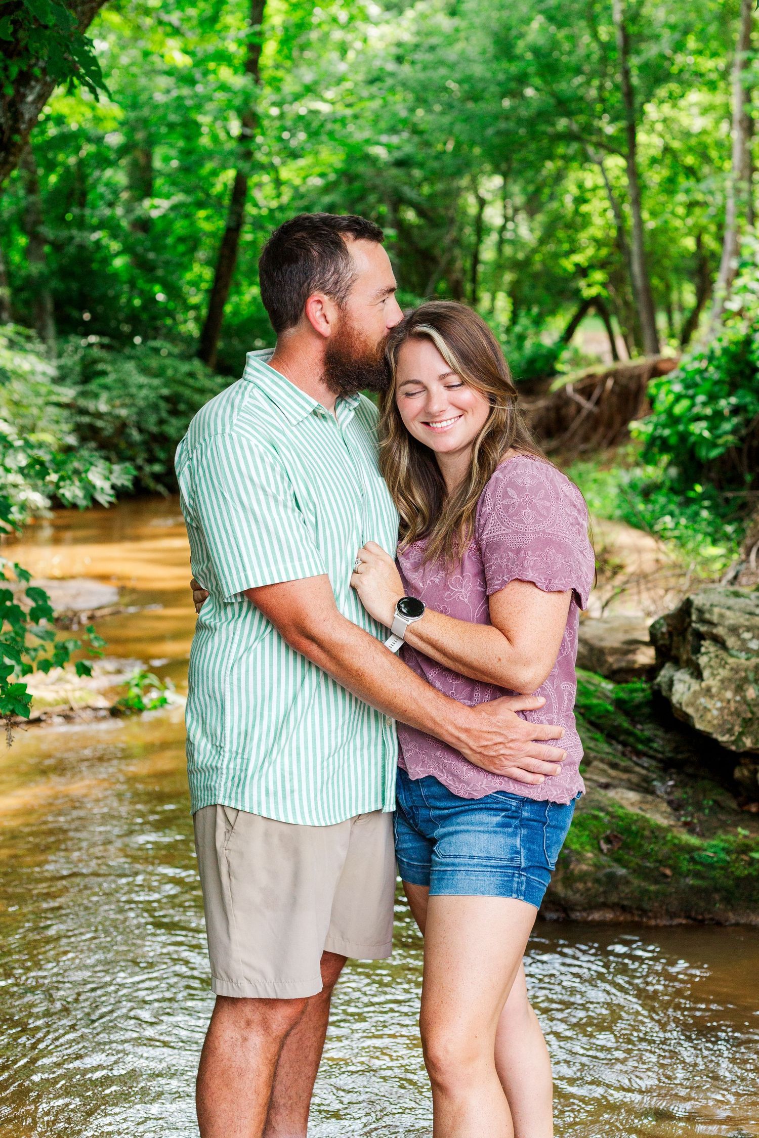 A couple embraces while standing in a sunlit creek with verdant forest surroundings.