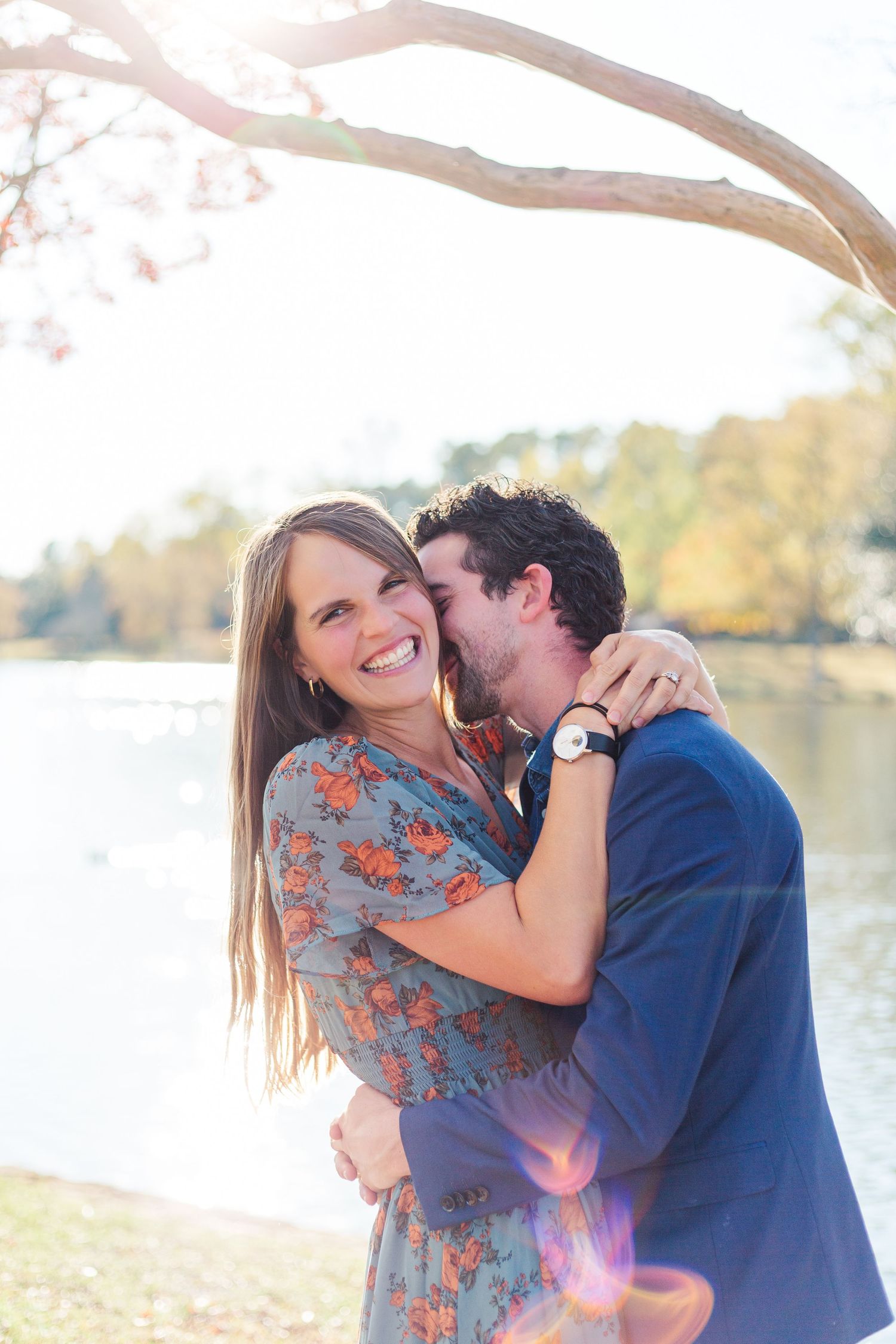 A romantic couple embraces by a lake during an autumn photo session with sunlight glowing through the trees.