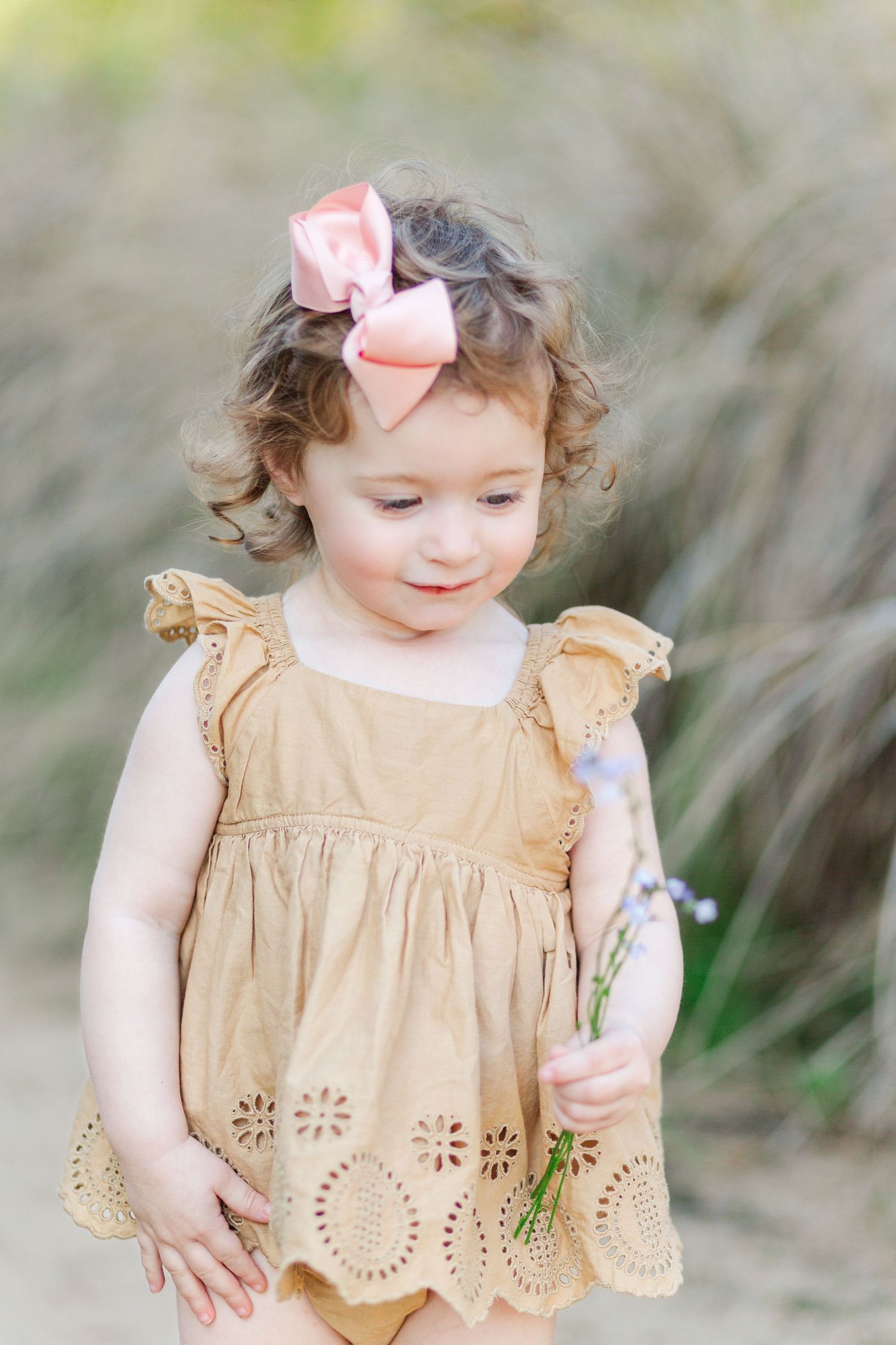 A toddler wearing a tan eyelet dress and pink bow plays outdoors in a natural setting.