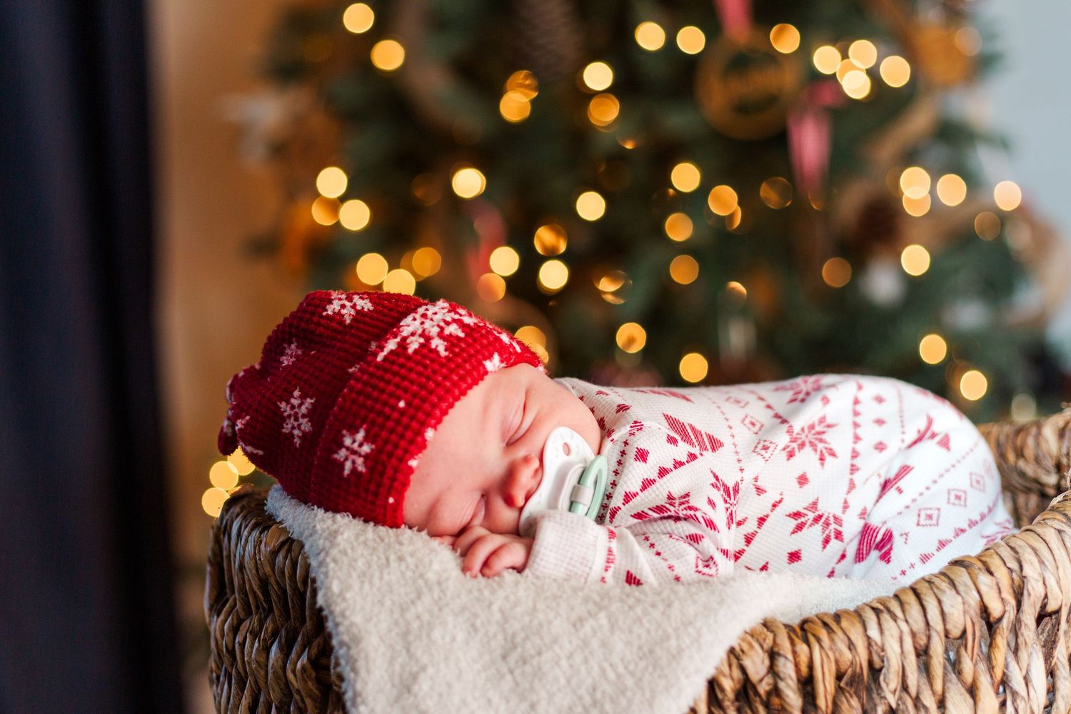 Sleeping baby in red snowflake pajamas rests in a wicker basket in front of a Christmas tree with twinkling lights.