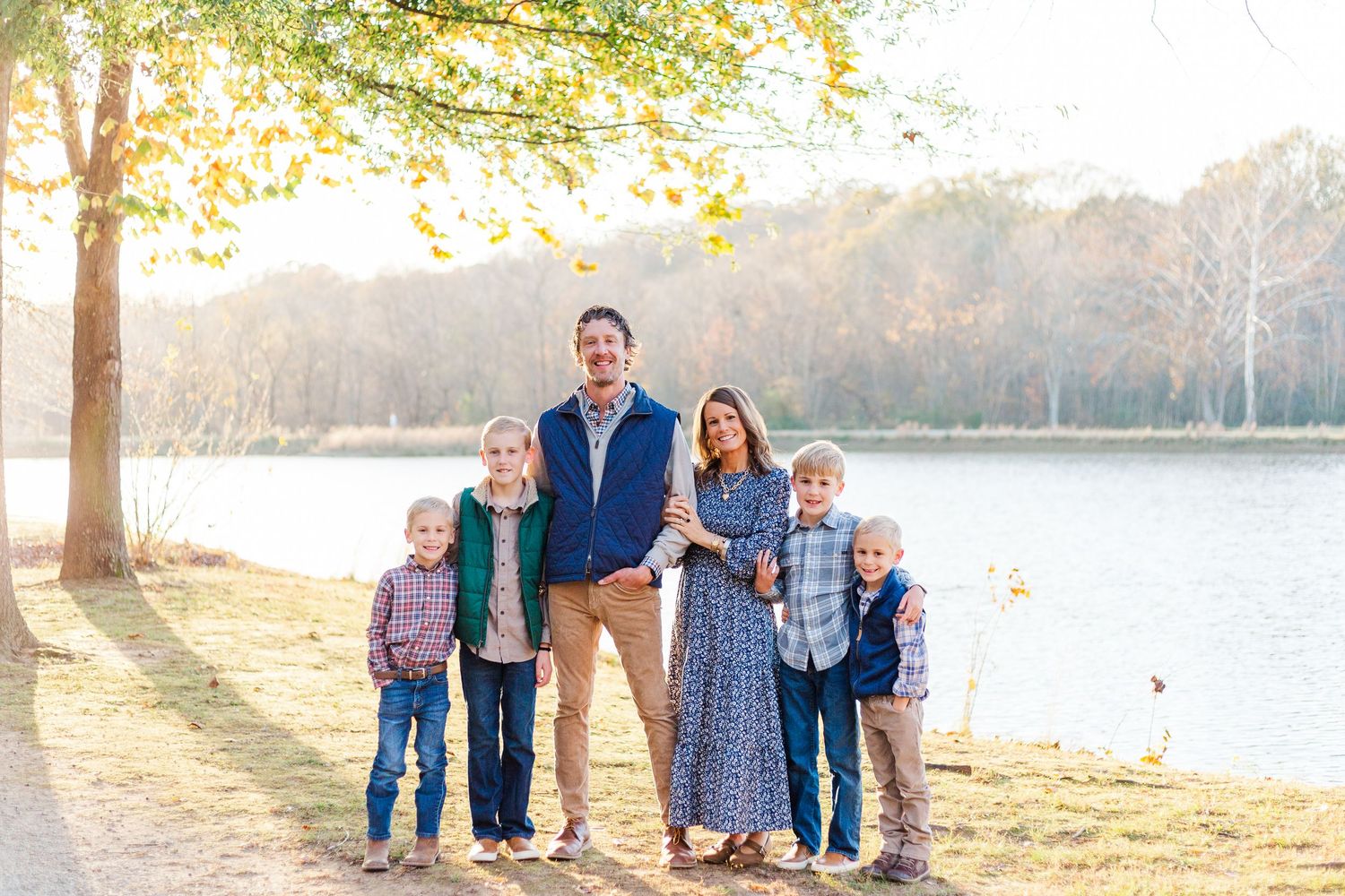 Family enjoying autumn sunset by lake with golden sunlight filtering through trees and fall foliage in background.