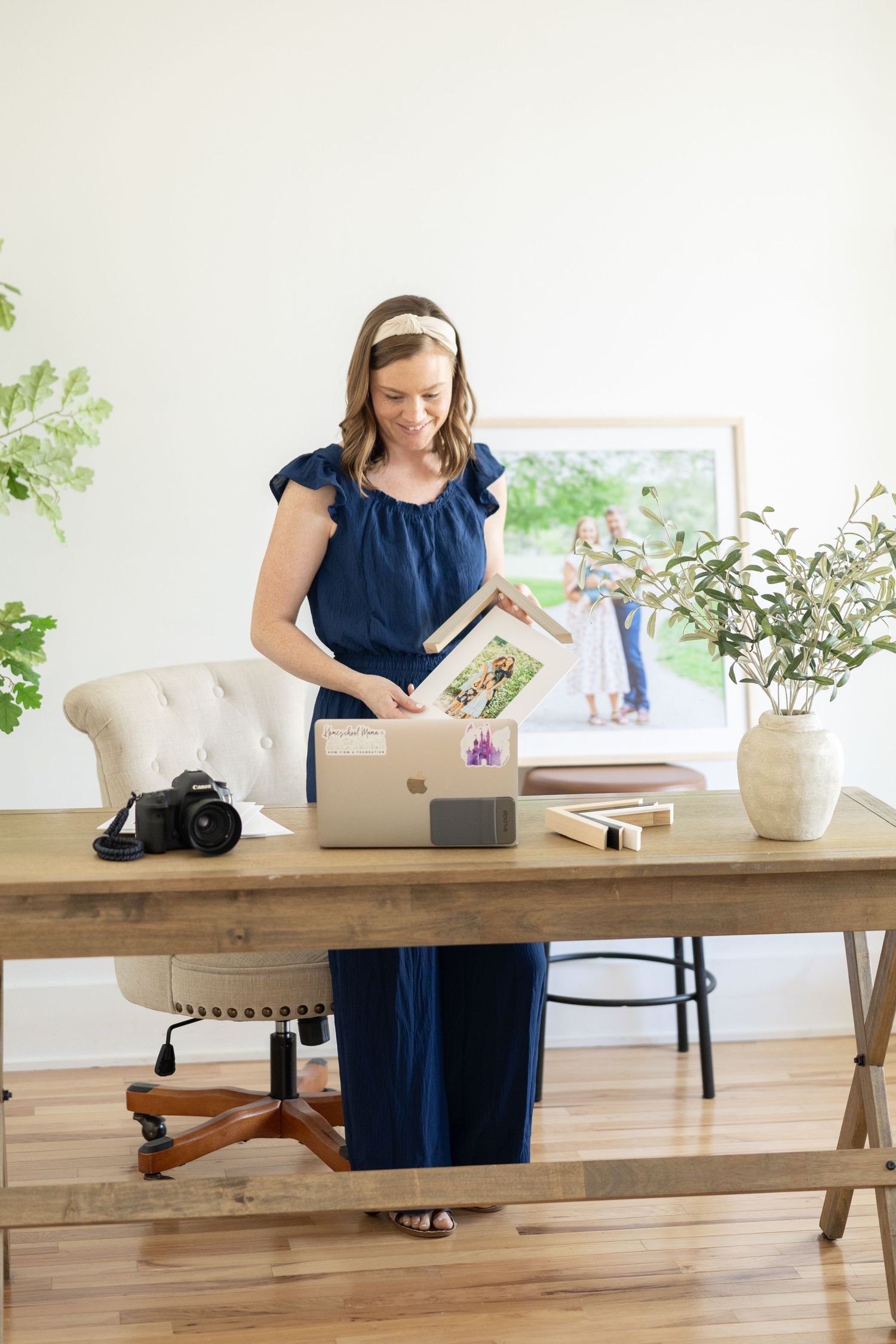 Person in navy blue dress working at modern wooden desk with laptop and plants in bright minimalist room.