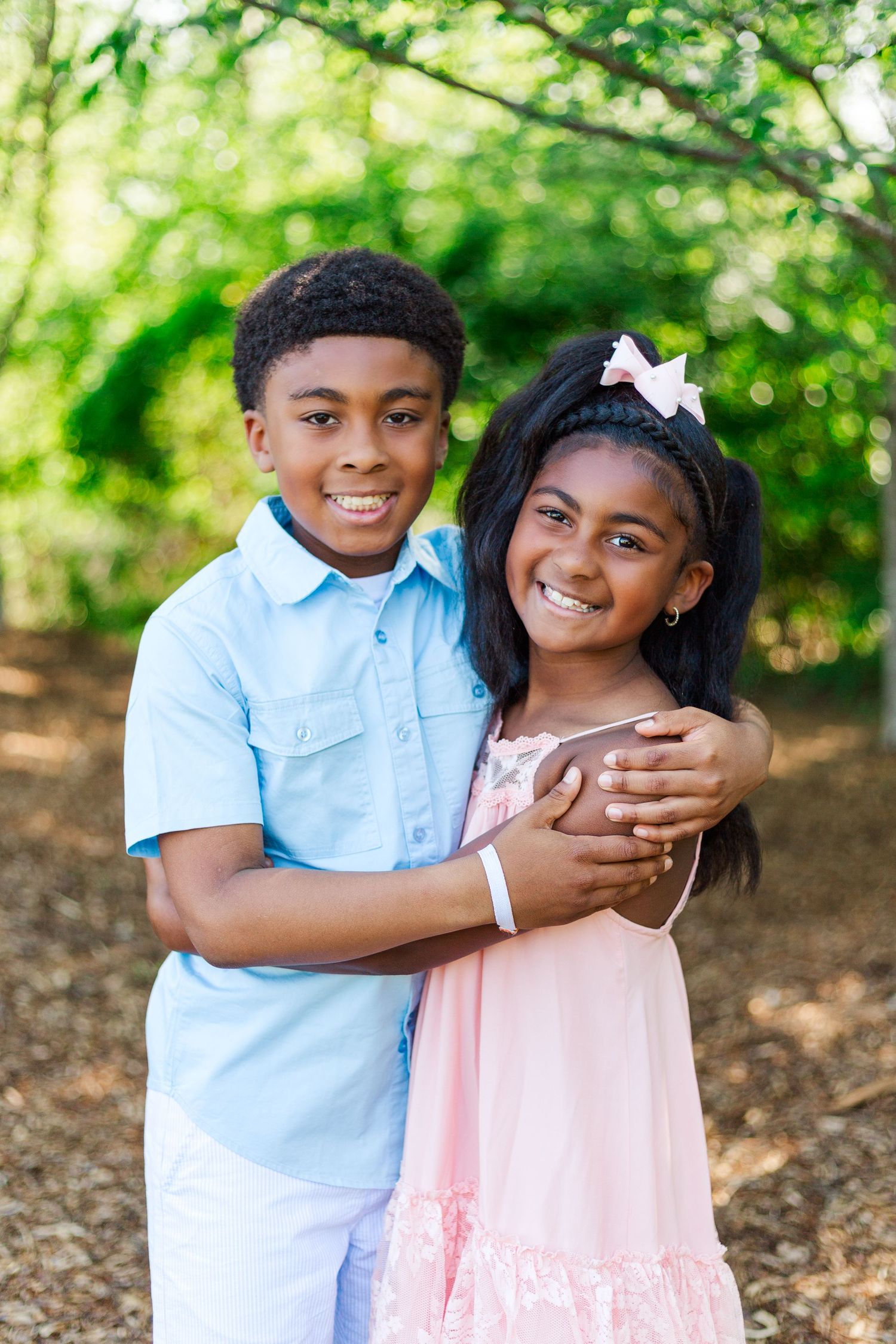 Two smiling children hug outdoors, boy in blue shirt and girl in pink dress with bow.