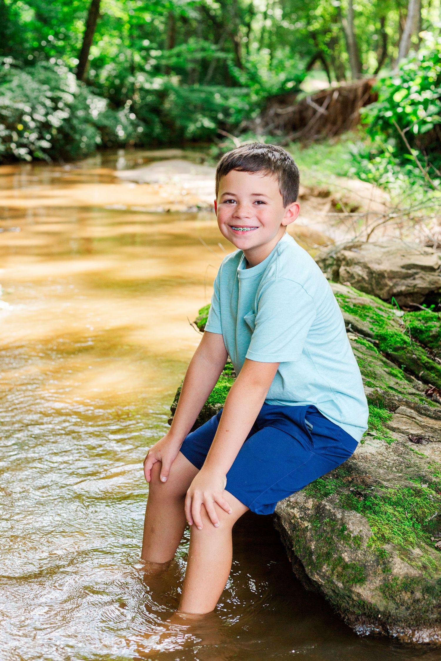 A child sits on a mossy rock by a peaceful creek in a lush forest setting.