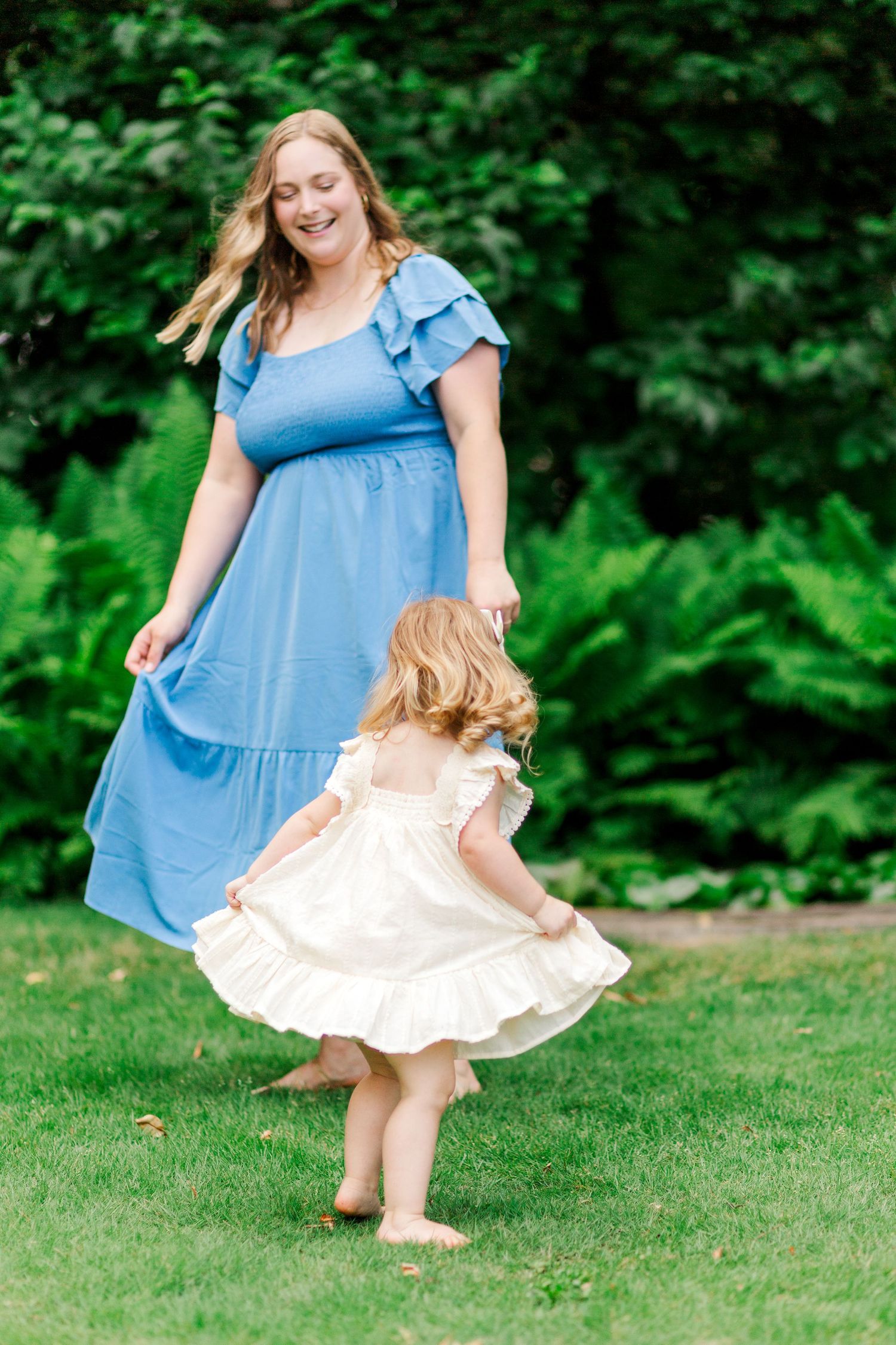 A mom and child in flowy dresses dance together in Clemson, SC joyfully on green grass surrounded by lush foliage for family photo session