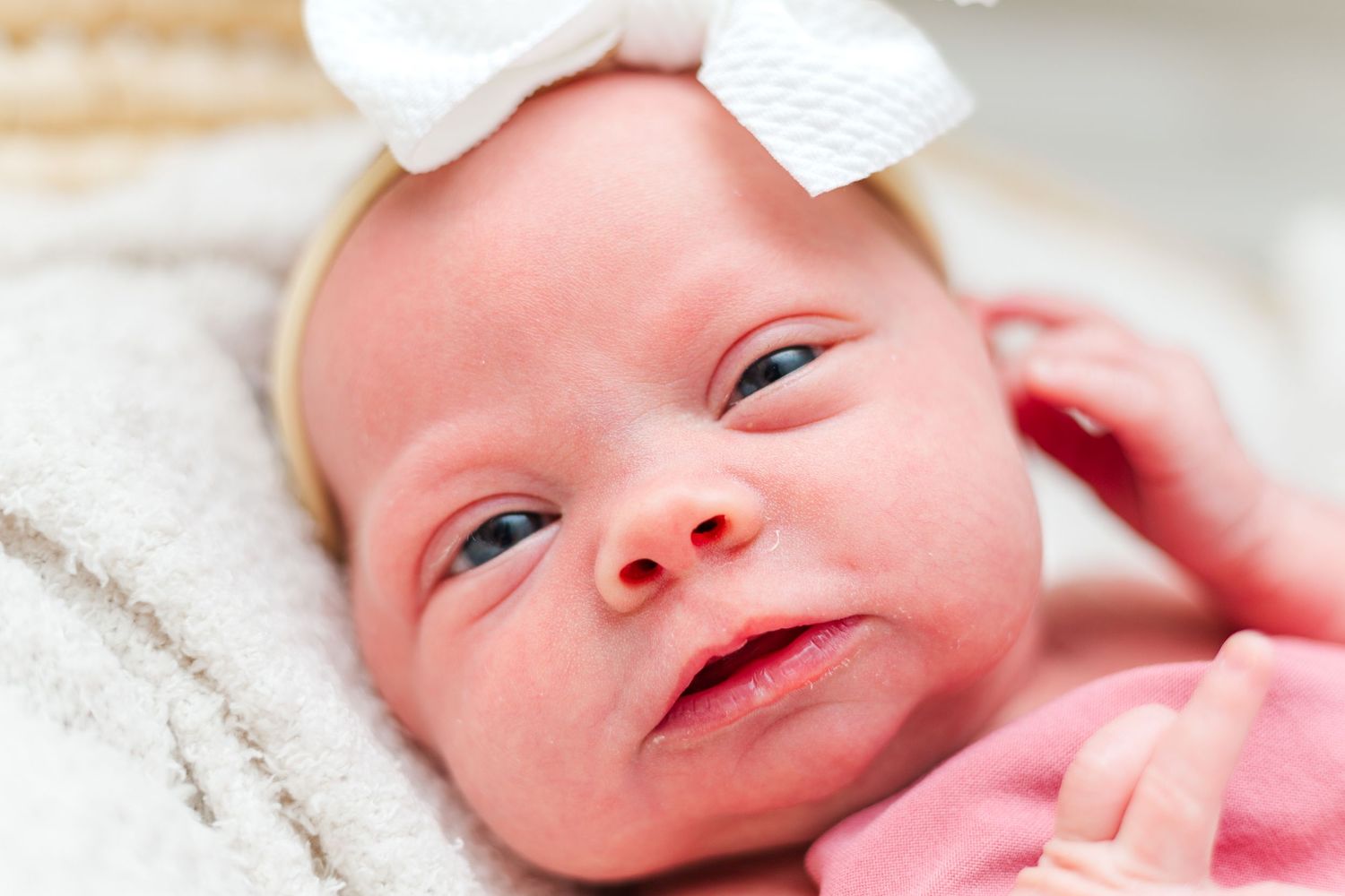 Sweet smiling infant wearing pink outfit and white headband rests on cozy white blanket.