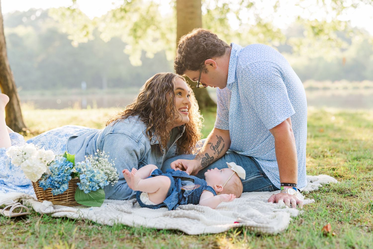 A young couple smiles at their baby on a blanket outdoors, surrounded by blue flowers in soft natural light.
