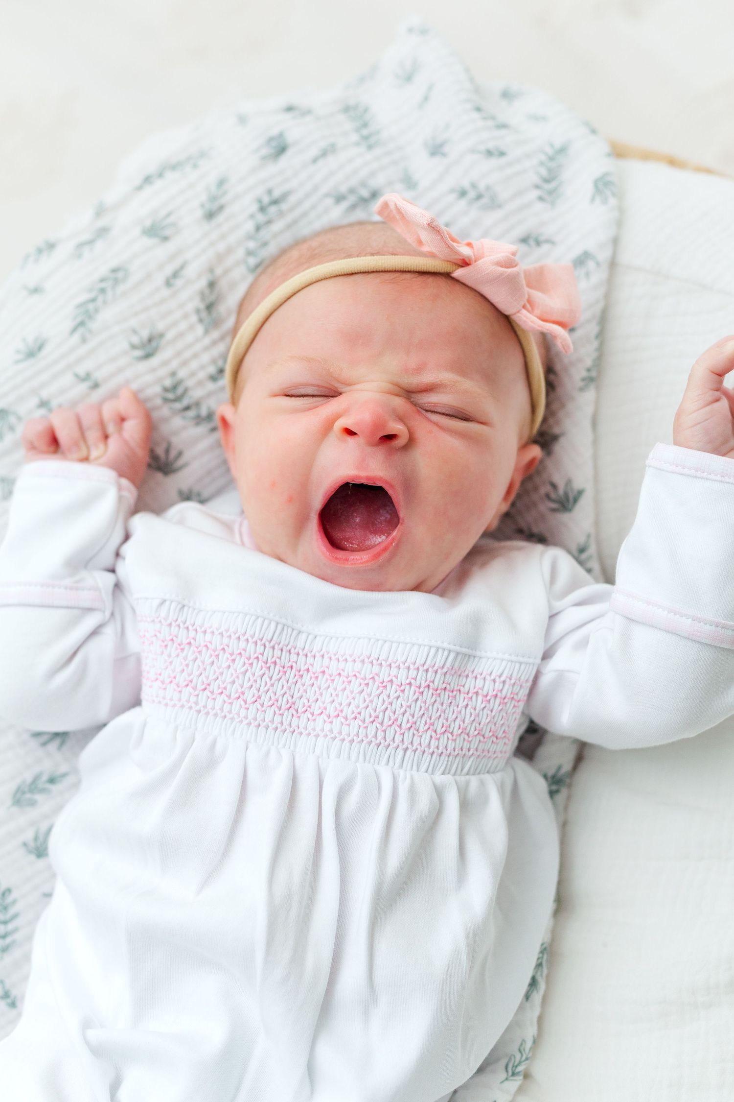 Newborn baby girl yawning in white smocked outfit with pink bow headband, lying on floral blanket.