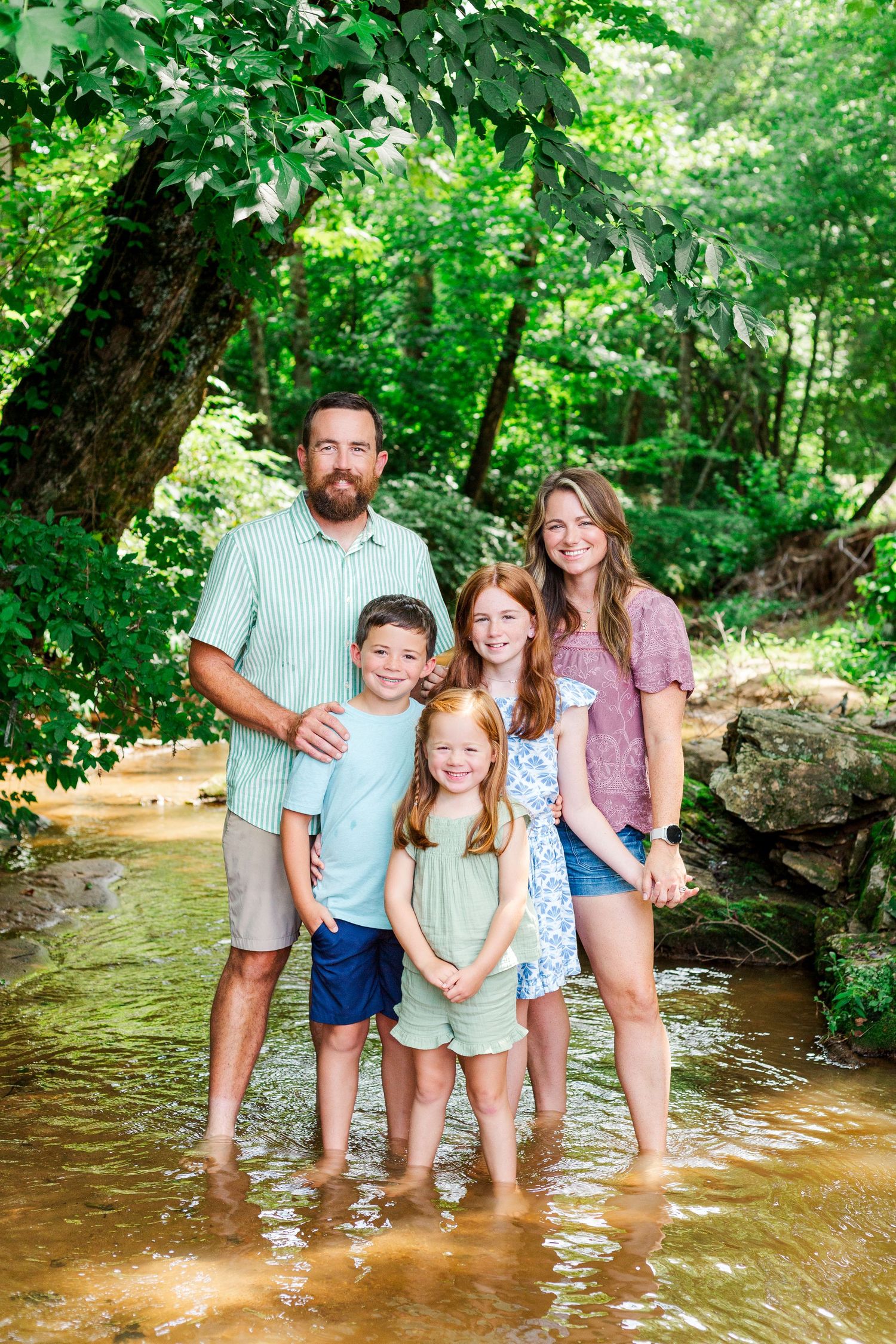 A cheerful family poses together while standing in a shallow creek with sunlit trees behind them.