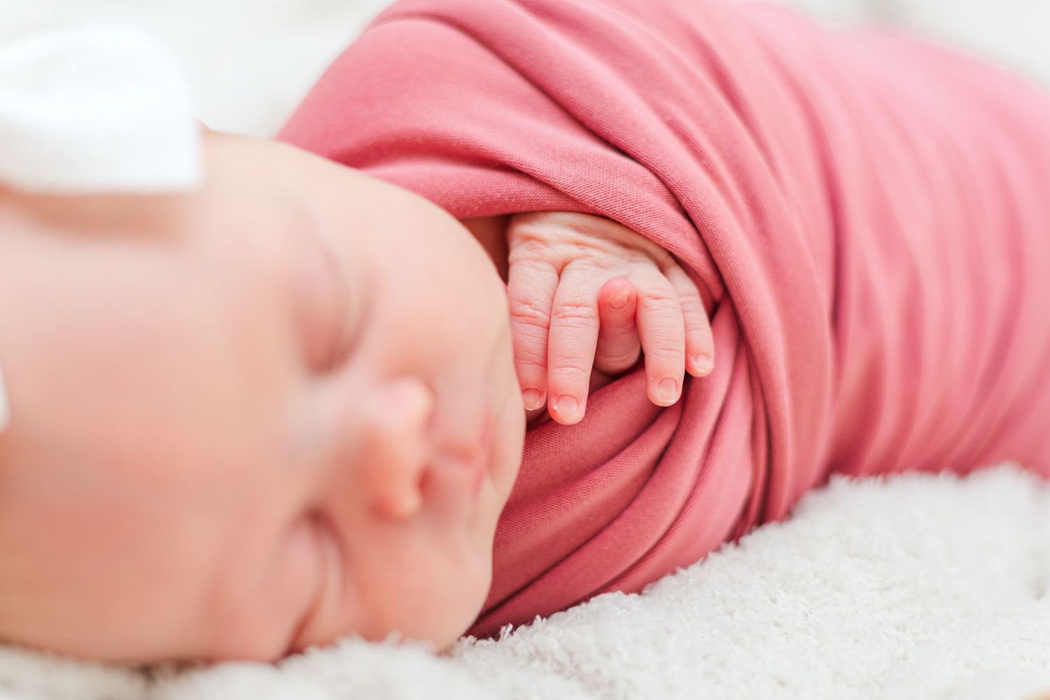 A newborn baby sleeps peacefully wrapped in a pink swaddle while their tiny hand rests gently on their face.