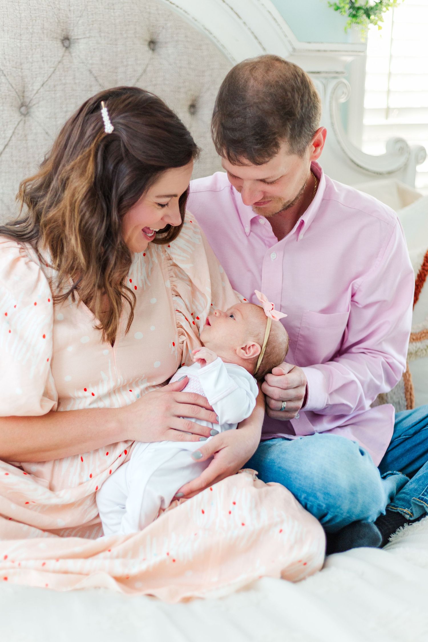 Happy parents sitting on bed smiling at their newborn baby girl in a pink bow headband.
