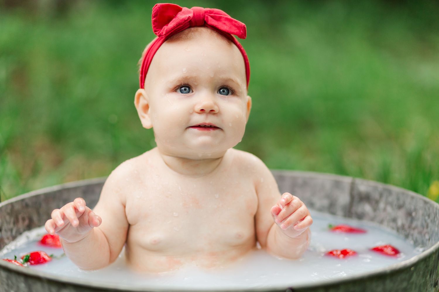 Baby wearing red headband sits in rustic wooden tub outdoors with green grass background in Greenville, SC for milestone session