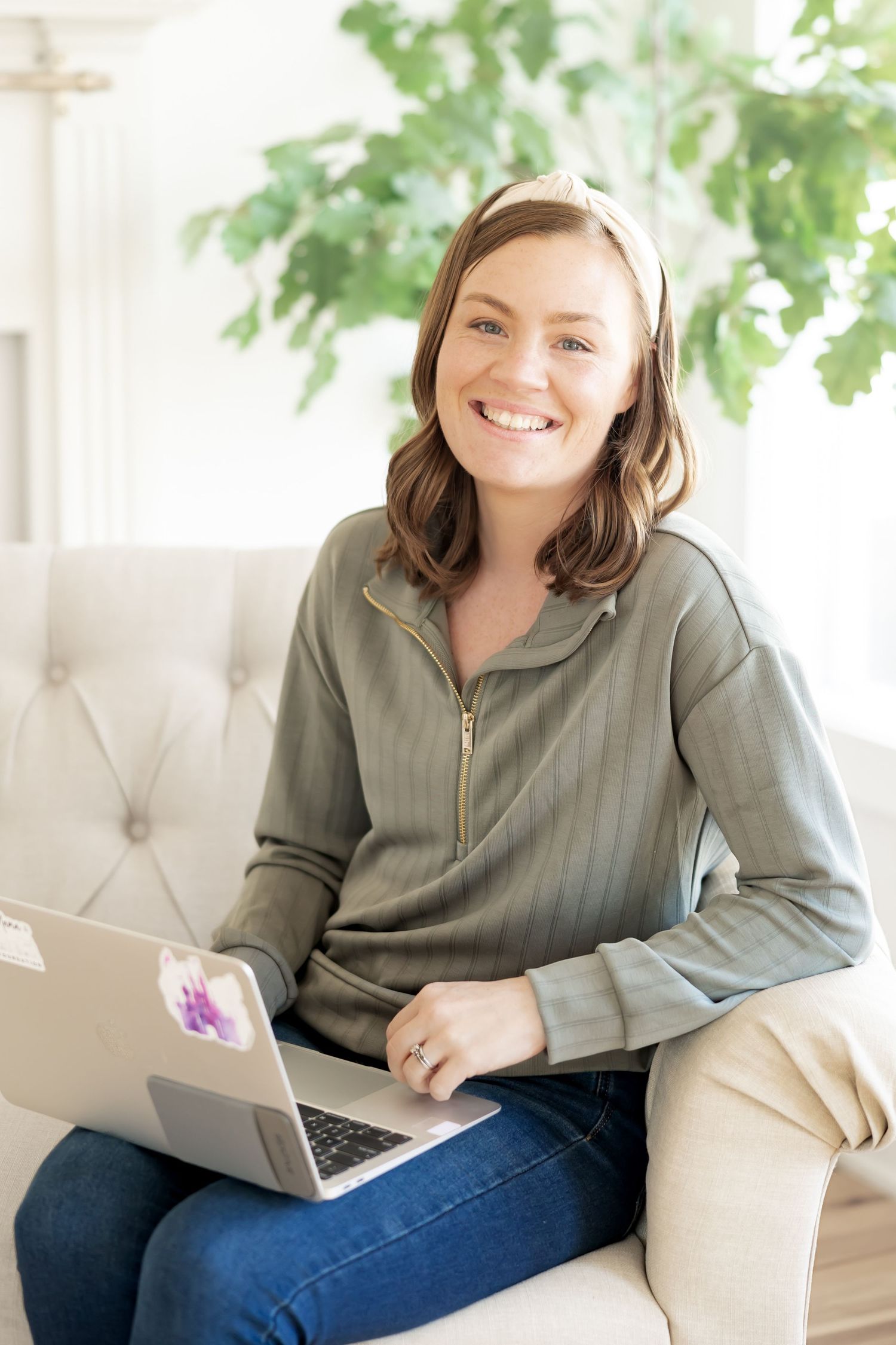 Smiling woman sitting on a sofa with a laptop, wearing a green zip-up top and jeans in a bright room.