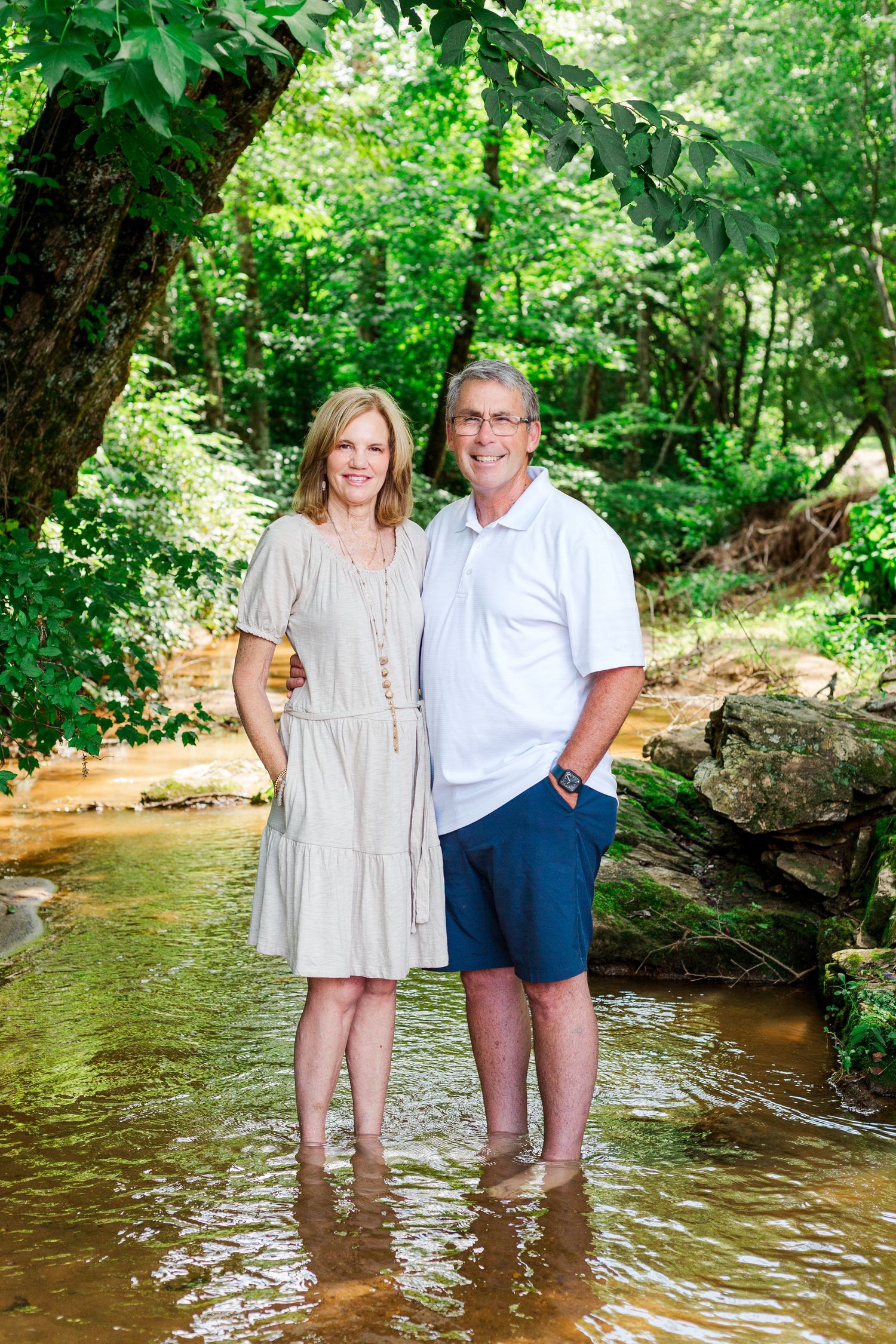 Two people stand together smiling in a stream surrounded by lush green trees and foliage.