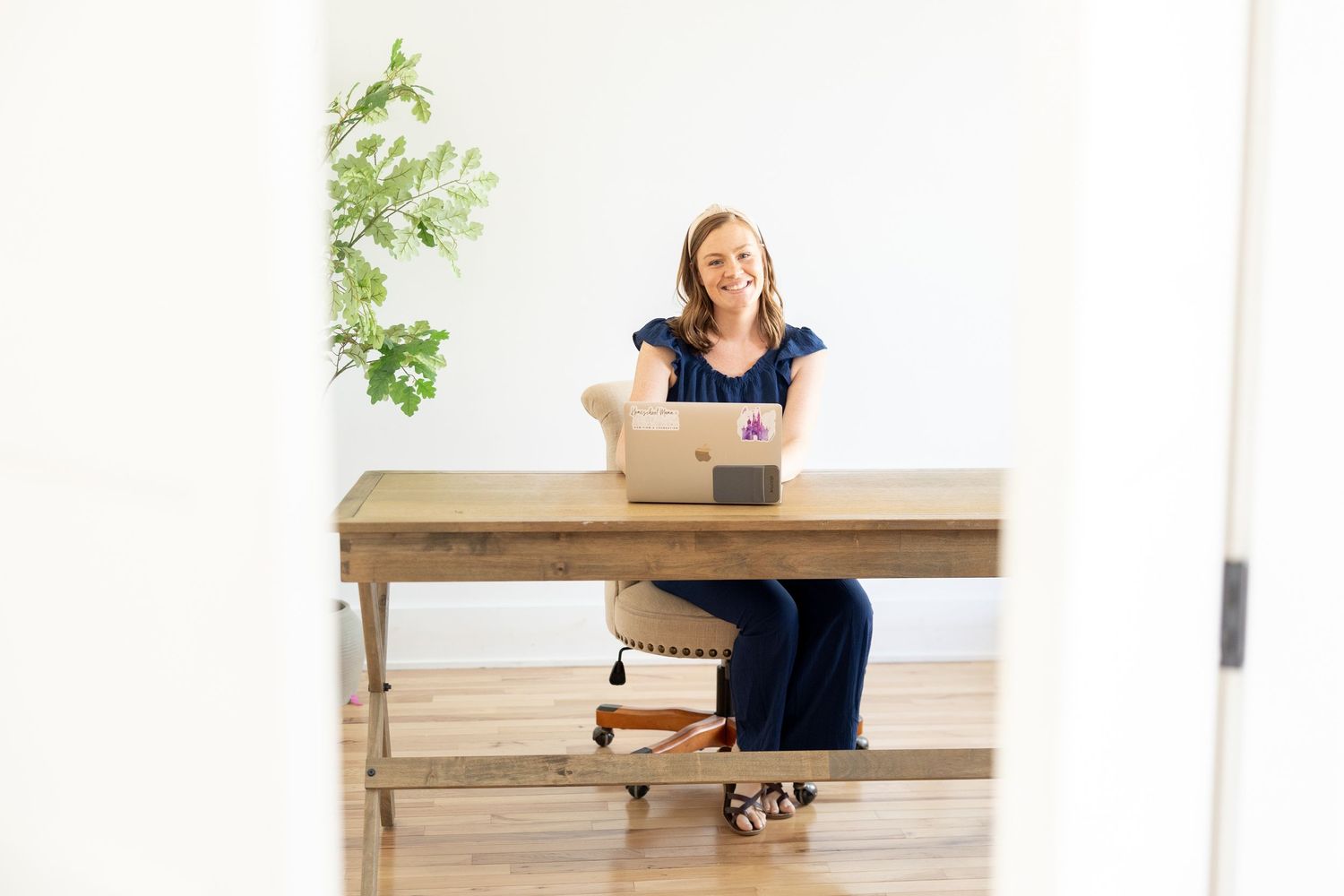Woman smiling at desk with laptop in bright modern home office with wood floors and green plant.