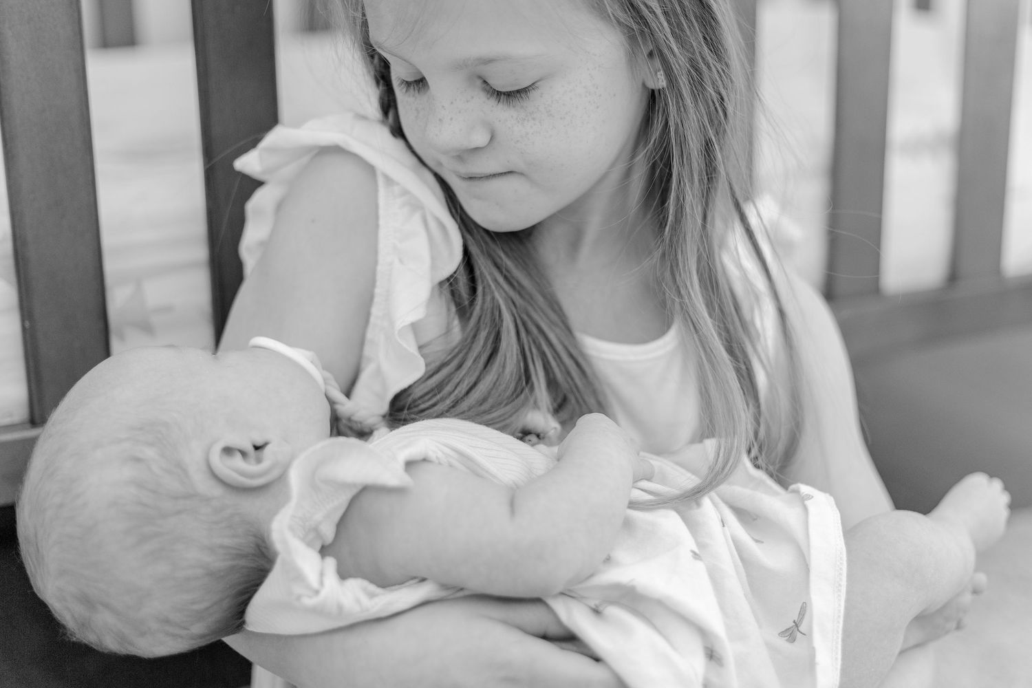A young child in a white shirt tenderly holds and gazes at a sleeping infant in a gentle black and white portrait.
