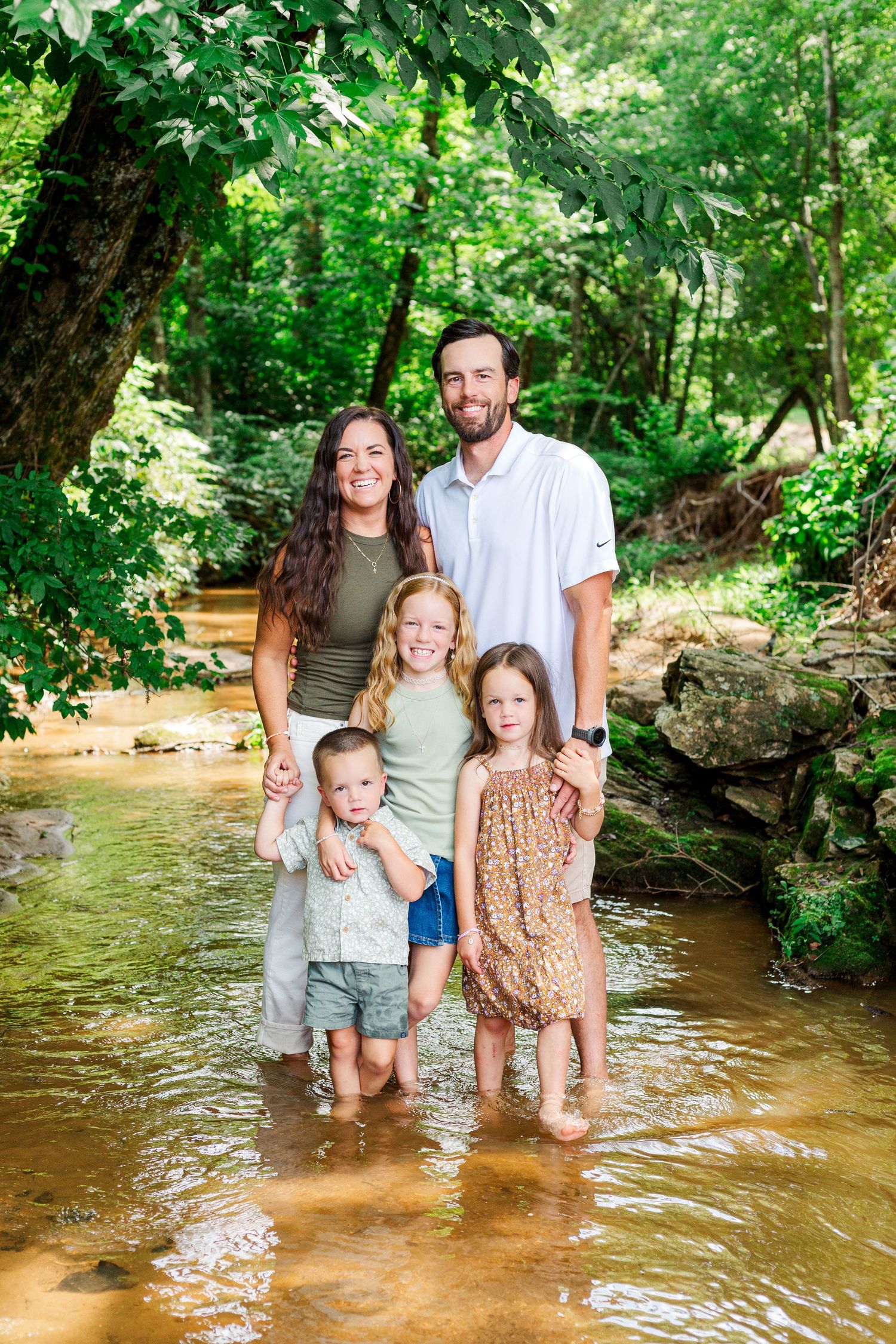 A family stands together in a shallow creek surrounded by lush green forest on a sunny summer day.