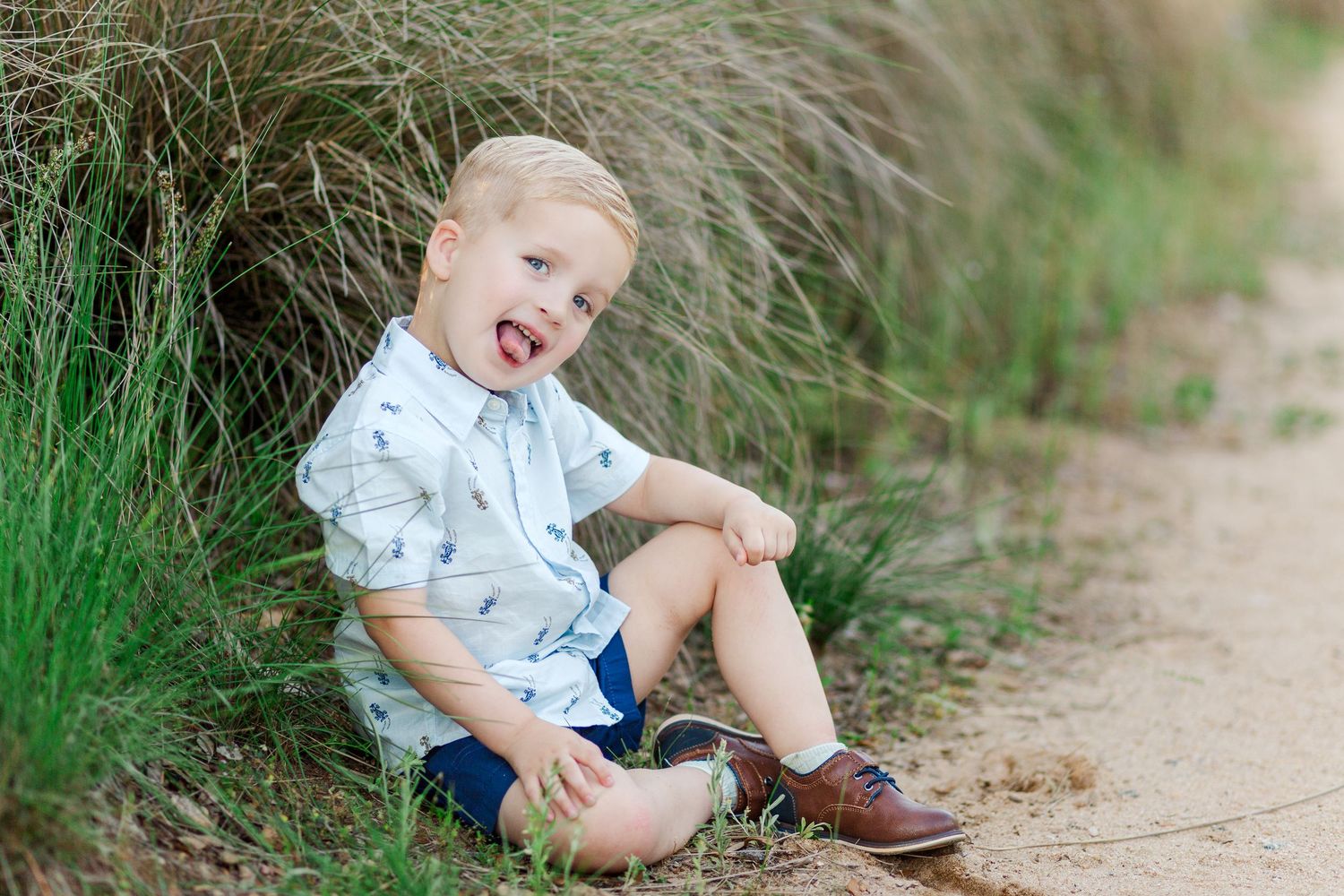 A child in blue sits on a dirt path surrounded by tall grass during a sunny outdoor photo session.