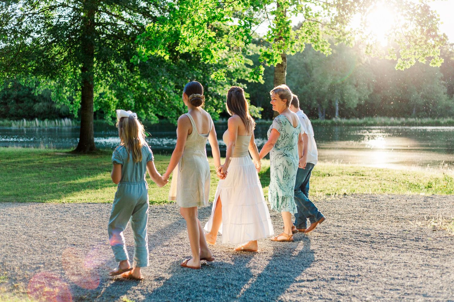 Family holding hands walking through a sunlit park near a pond surrounded by lush green trees.