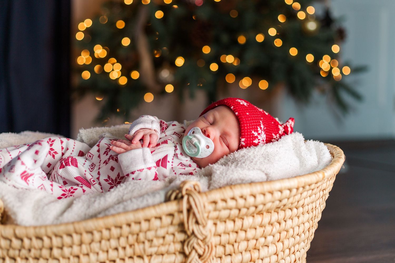 Baby wearing red santa hat lies swaddled in white blanket inside woven basket with Christmas tree lights bokeh in background.