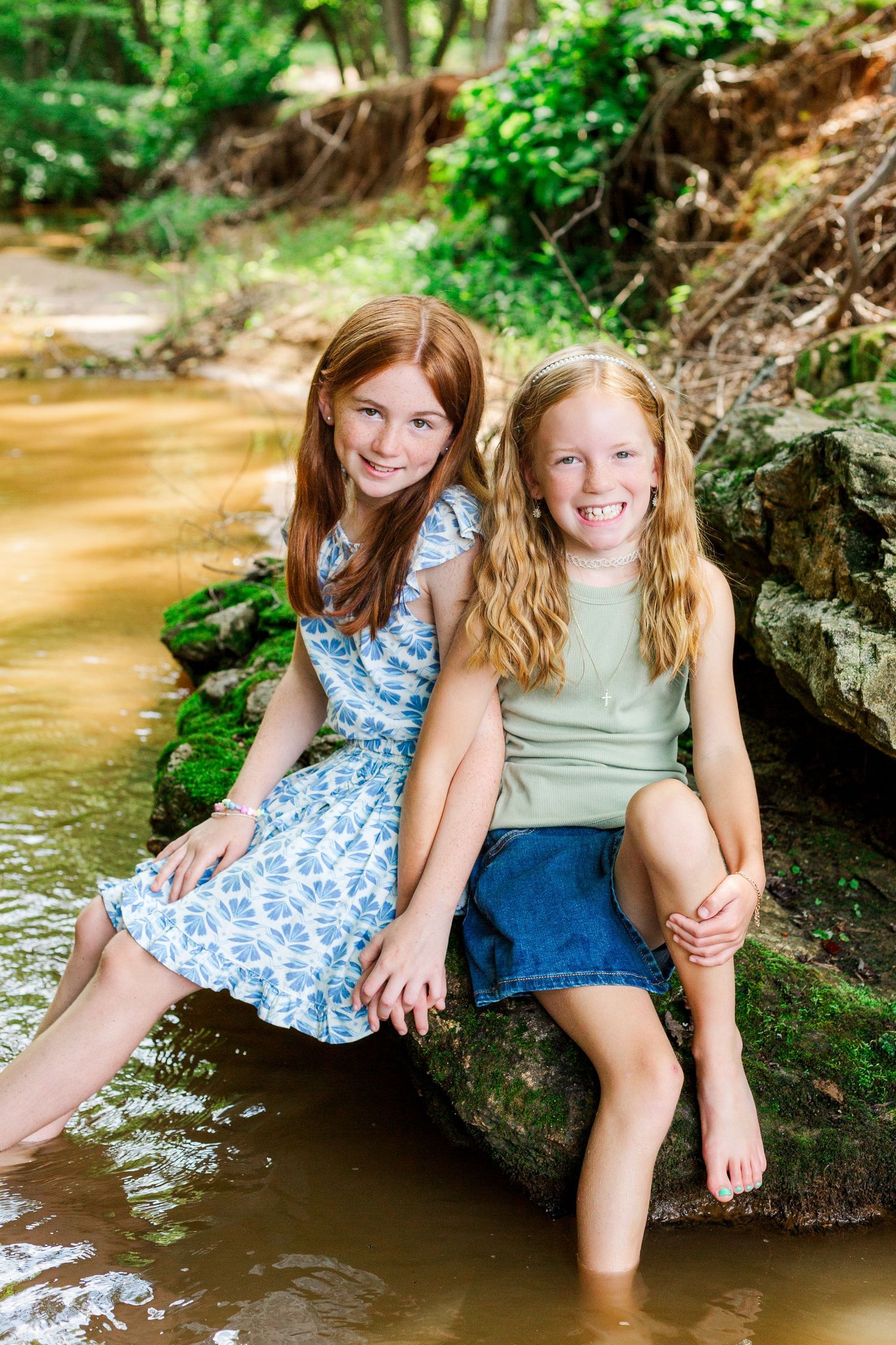 Two children sitting on rocks in a shallow creek surrounded by lush greenery on a bright day.