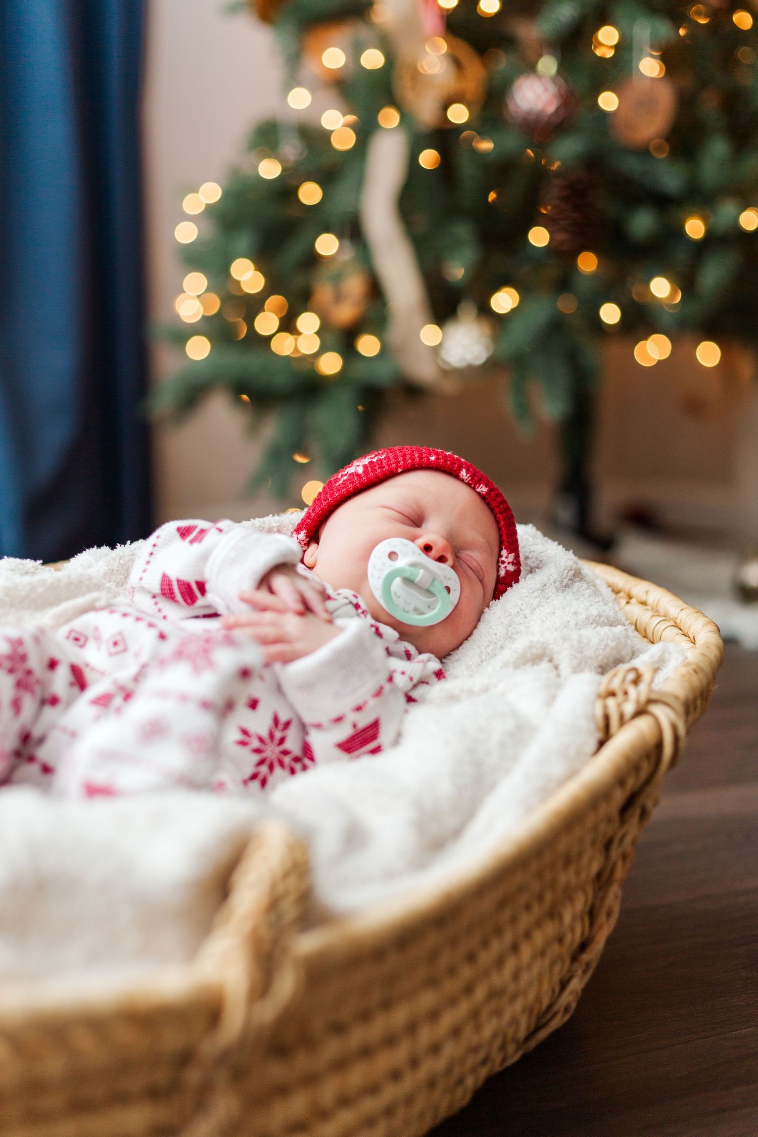 Sleeping infant wearing red hat rests in wicker basket with winter blanket under Christmas tree with glowing lights.