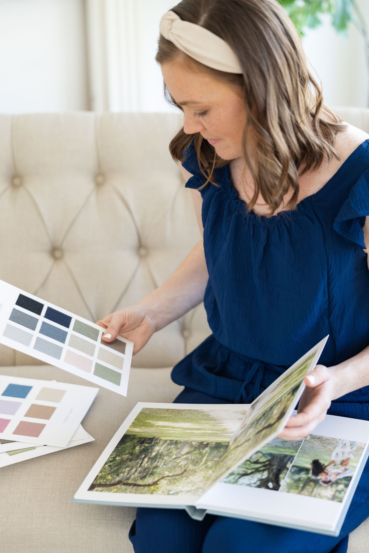 Woman in navy dress reviewing color swatches and photo album on beige tufted sofa.