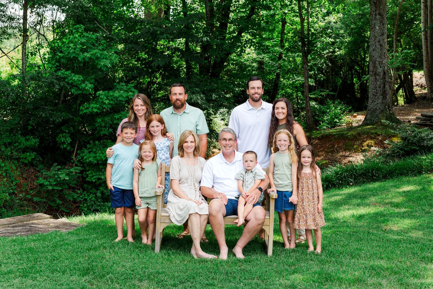 Group of adults and children in coordinated light-colored outfits pose together on a grassy lawn with lush trees in background.