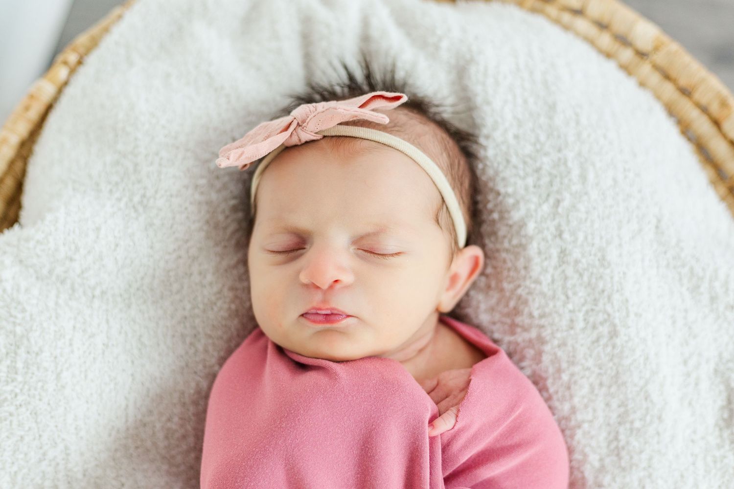 Sleeping newborn baby girl wrapped in pink swaddle with bow headband resting in a wicker basket.