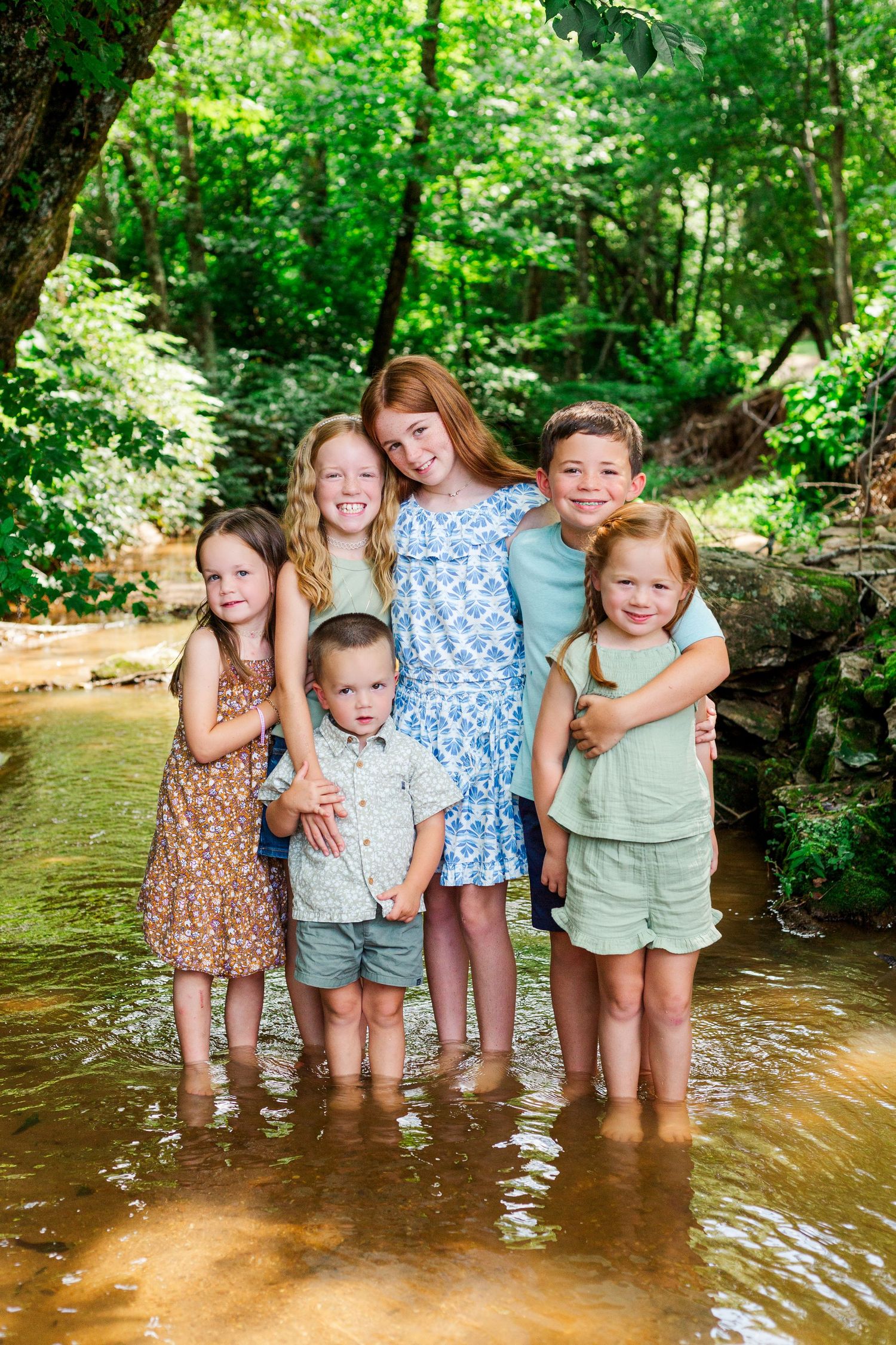 Group of children standing together in a shallow creek surrounded by lush green forest on a sunny summer day.