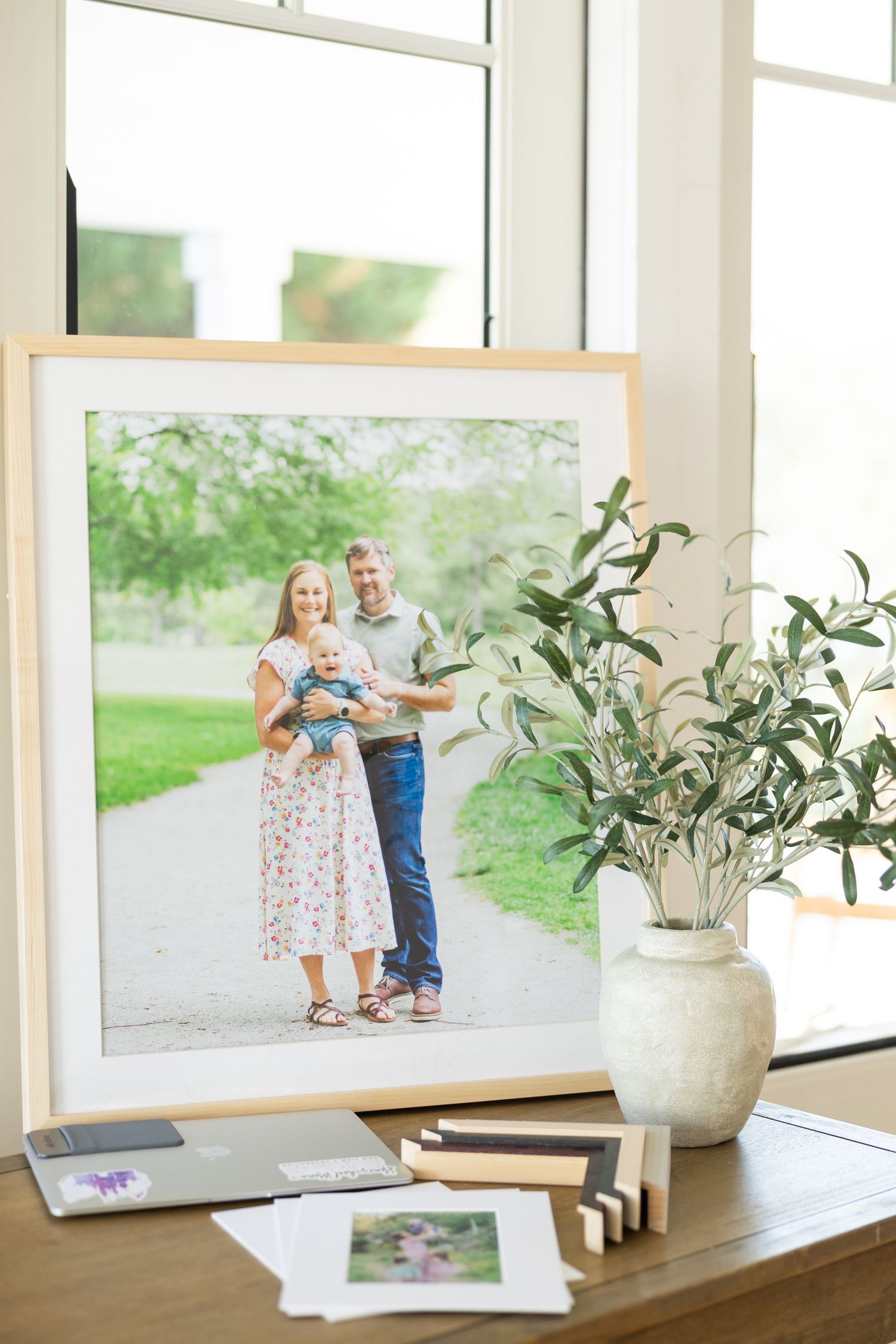 A framed family photograph sits on a wooden table with a ceramic vase of olive branches and scattered prints nearby.