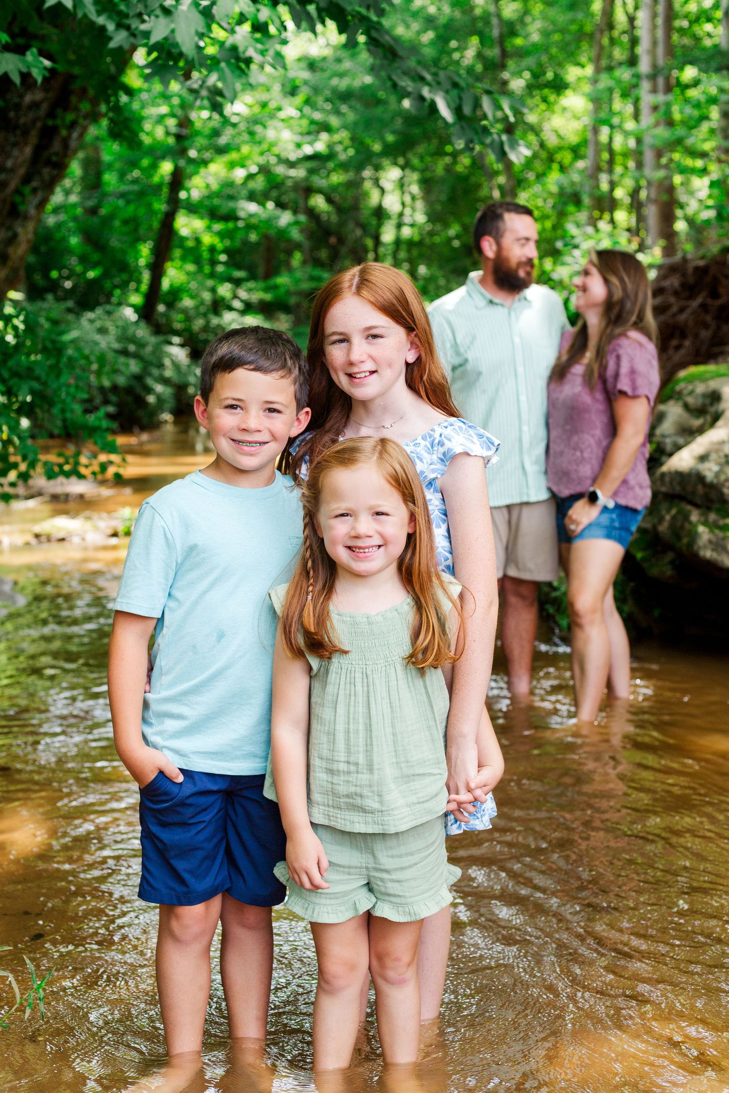 Young children stand in shallow creek water while families gather in the background.
