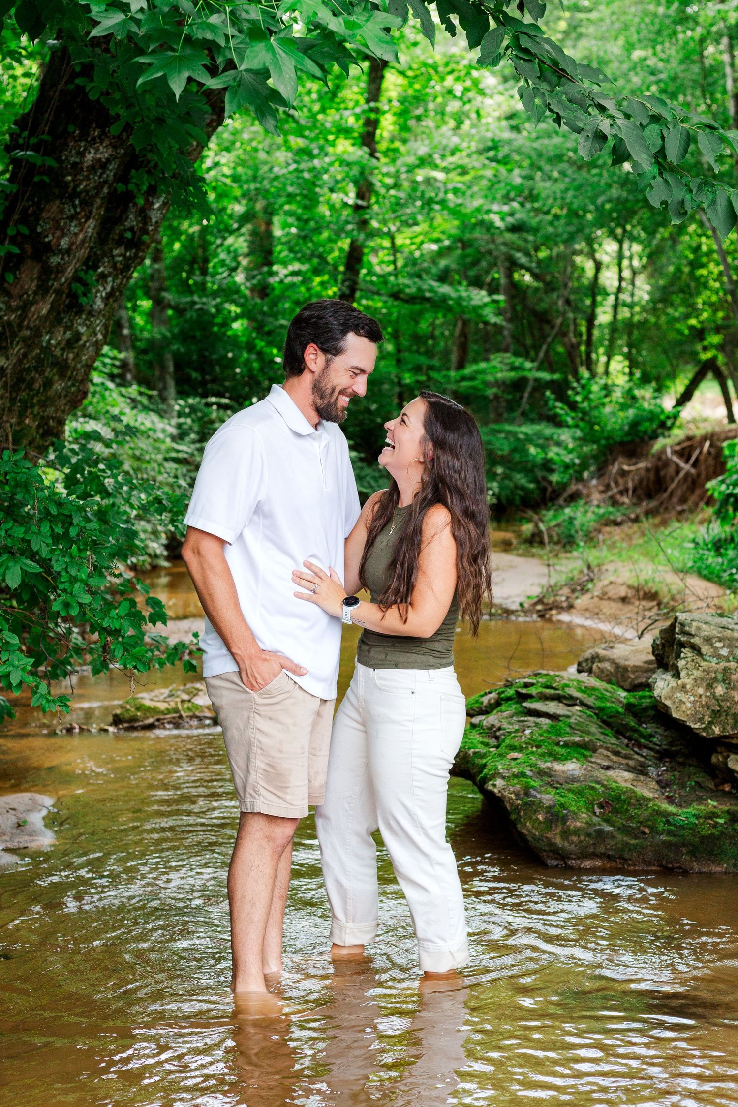A couple shares a tender moment while standing barefoot in a shallow creek surrounded by green foliage.