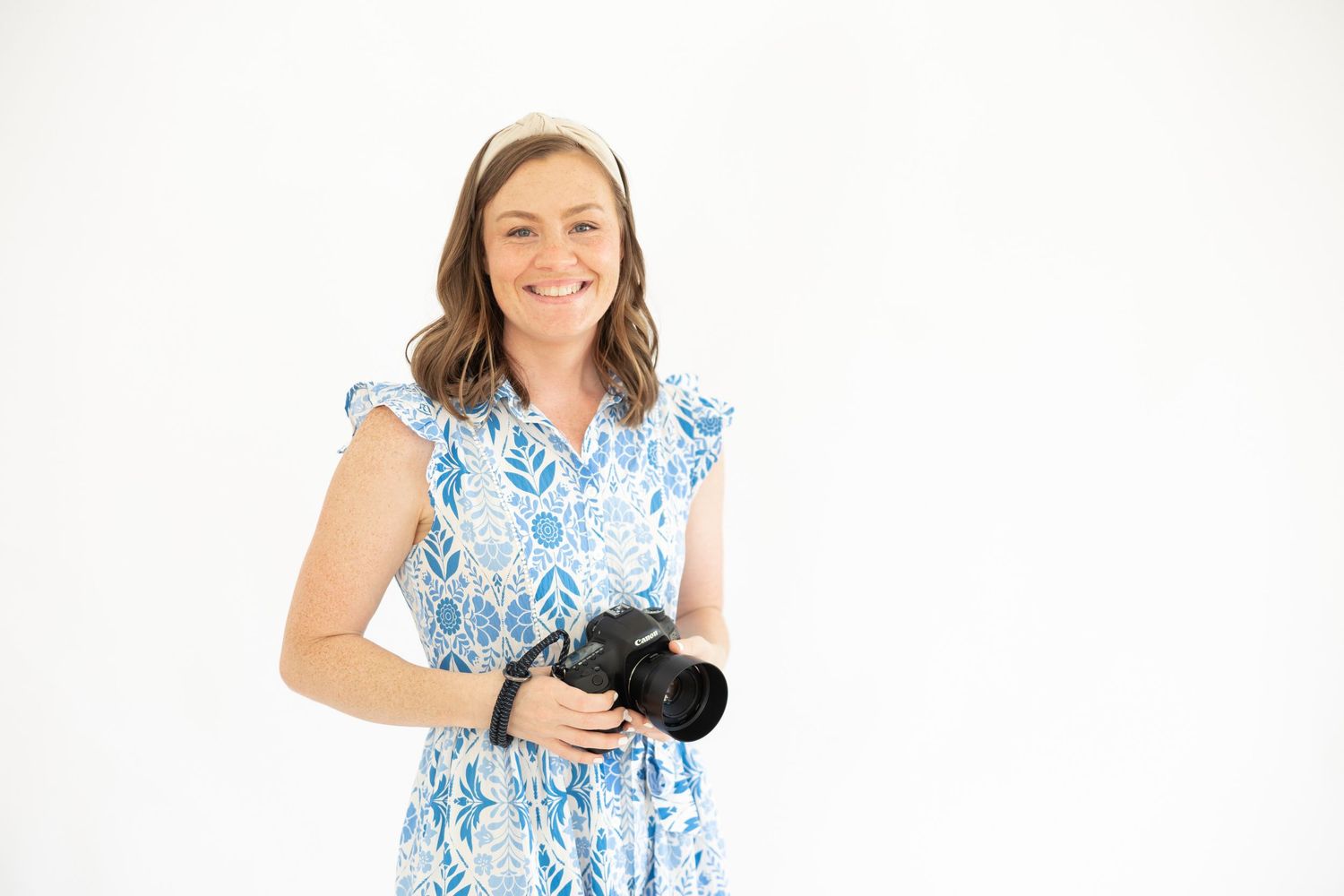 Smiling woman in blue floral dress holding a DSLR camera, posing against a white background.
