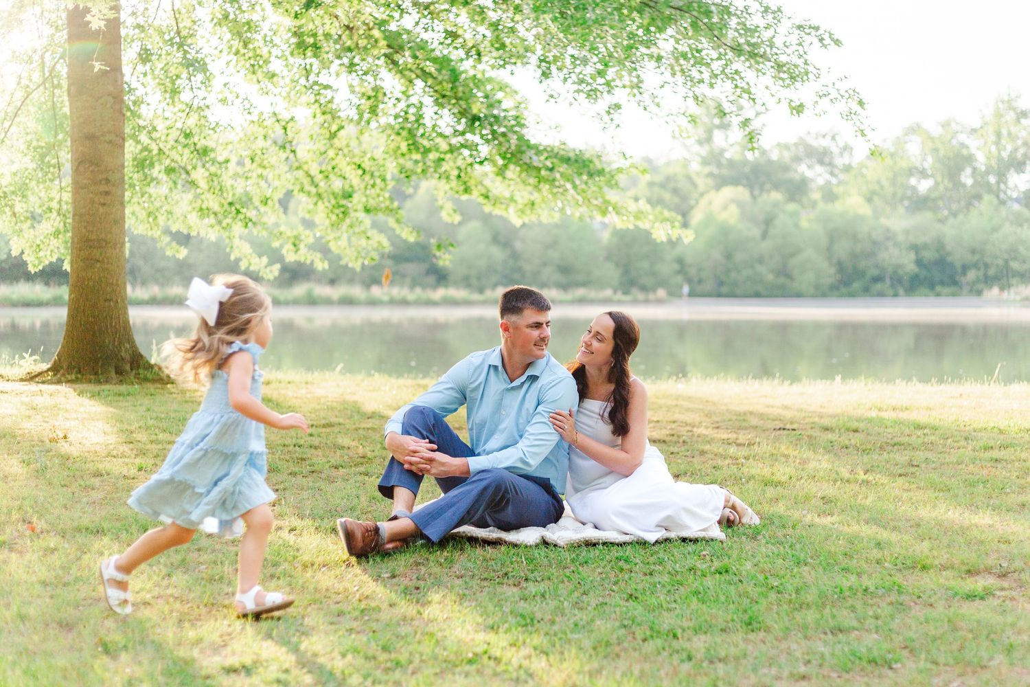 Family sitting on grass by a lake, with a young girl in a blue dress running toward her parents on a sunny day.
