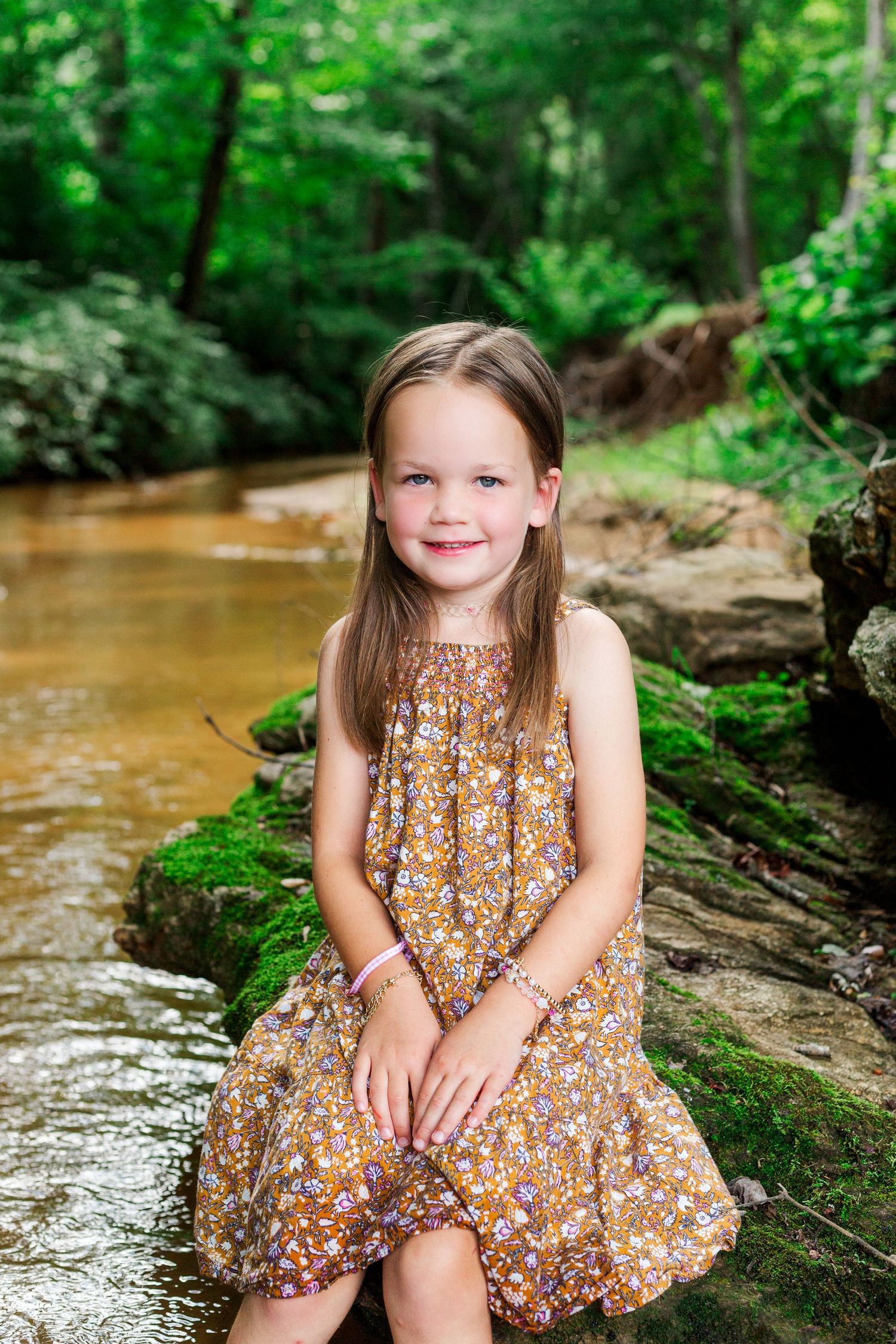 A floral dress adorned subject sits on a mossy rock by a flowing stream in a wooded setting.