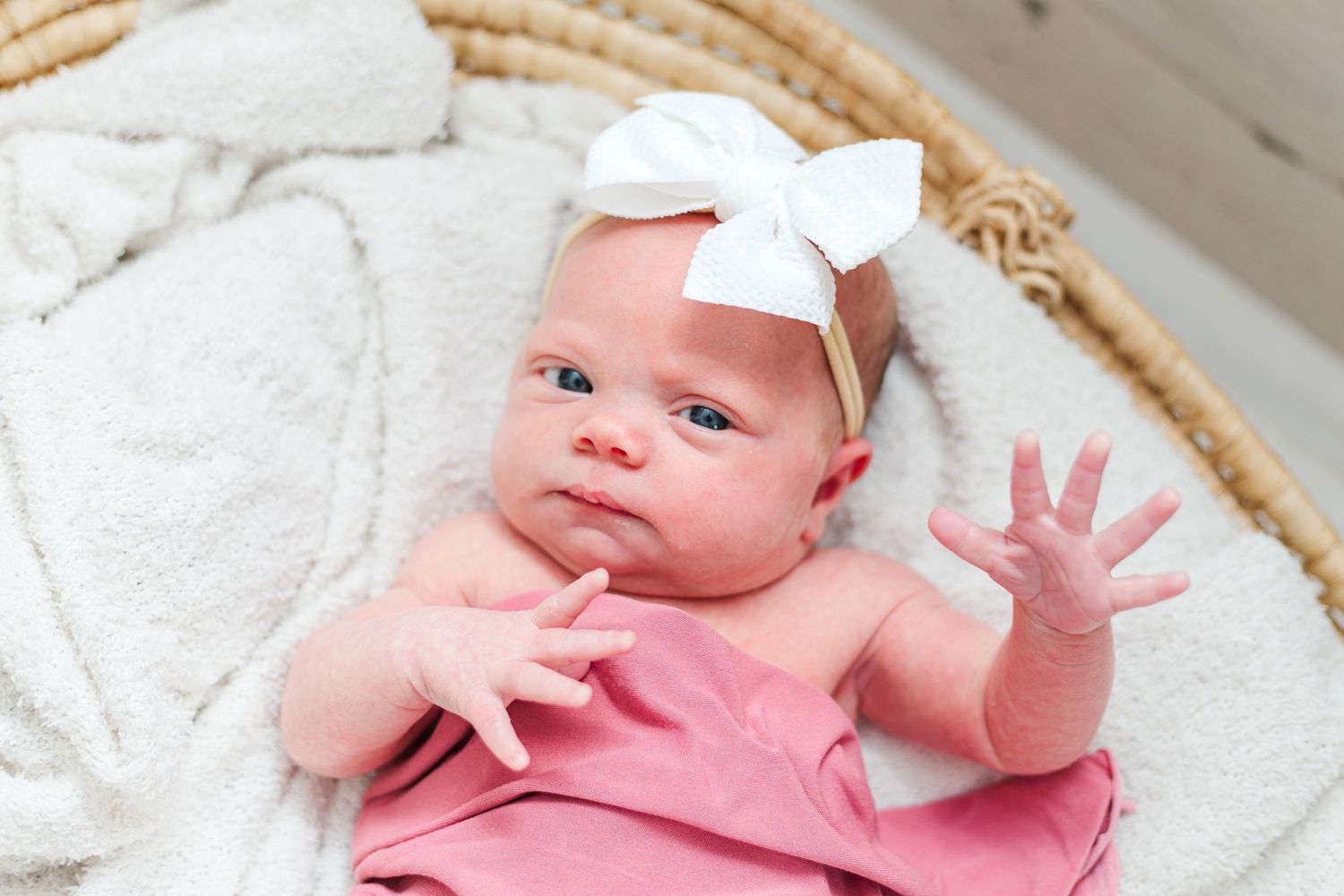 Infant in pink shirt lying on white blanket in woven basket smiles lovingly at camera.