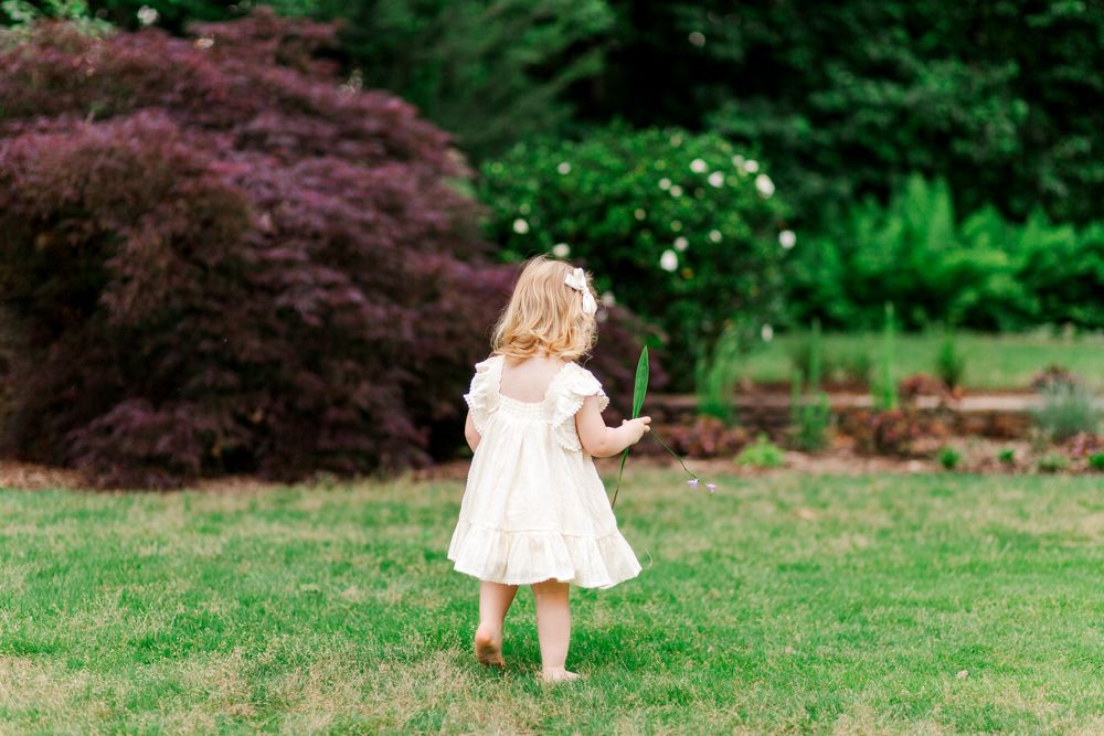 Small child in white dress running across a lush green lawn with flowering trees and bushes in the background.