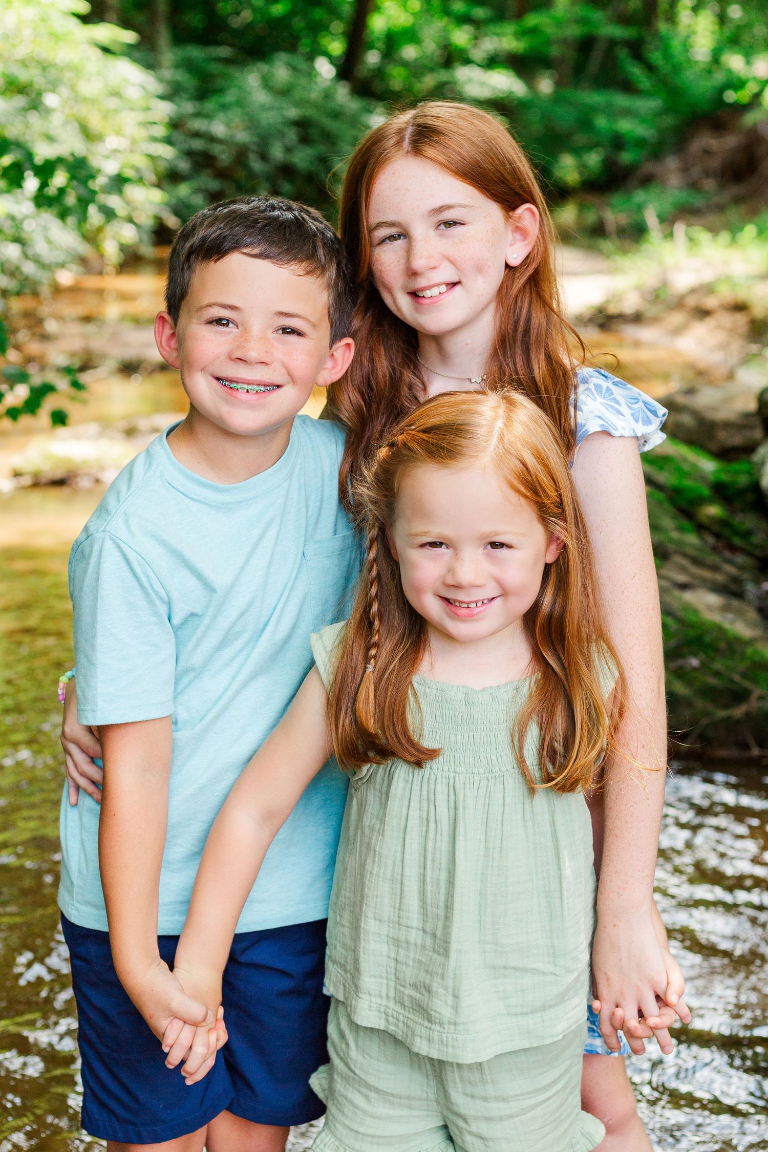 Three children stand close together in a creek with green foliage in the background.