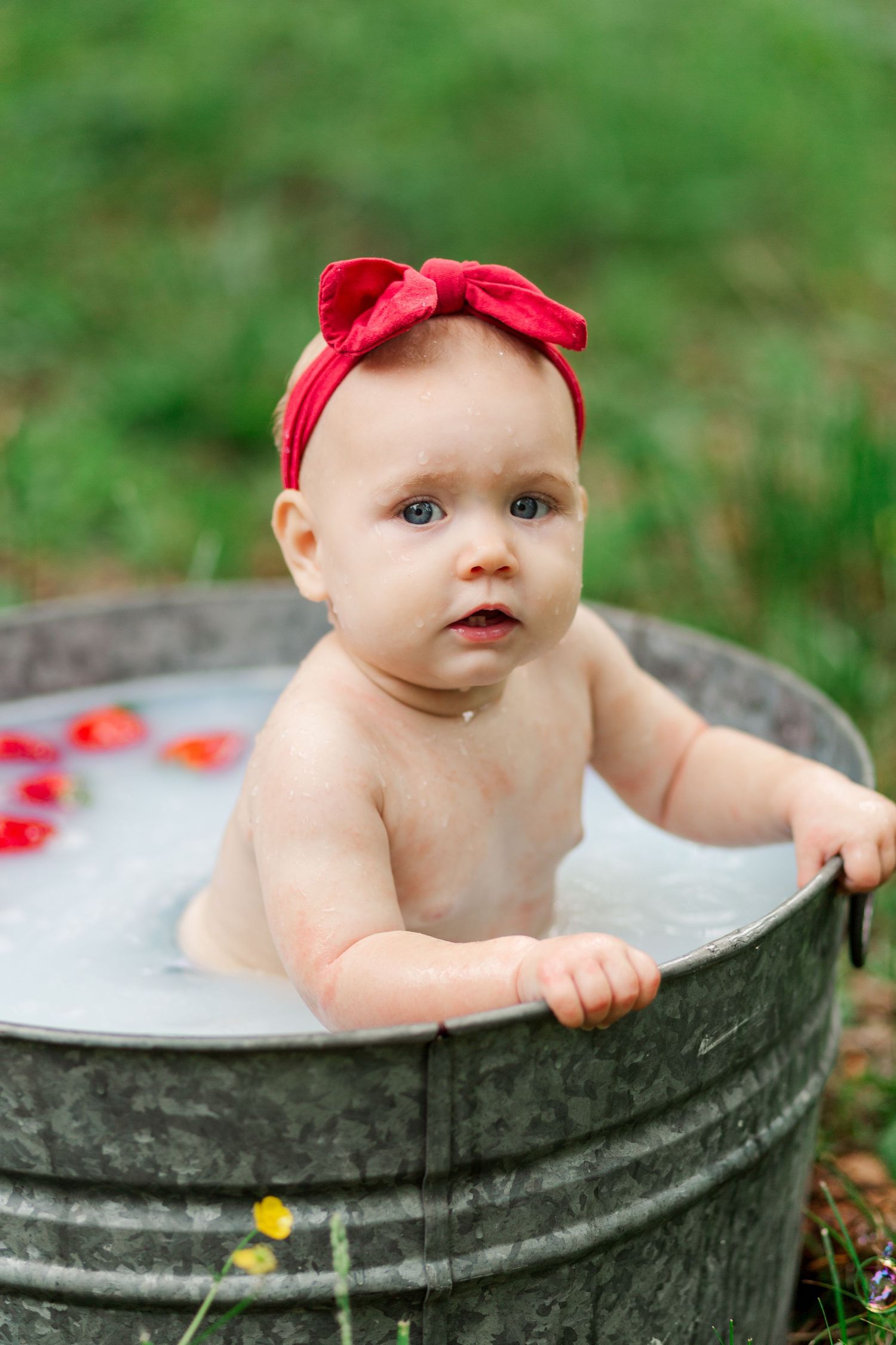 A baby wearing a red bow sits in a rustic metal washtub for a charming outdoor photo session in Greenville, SC for milestone session