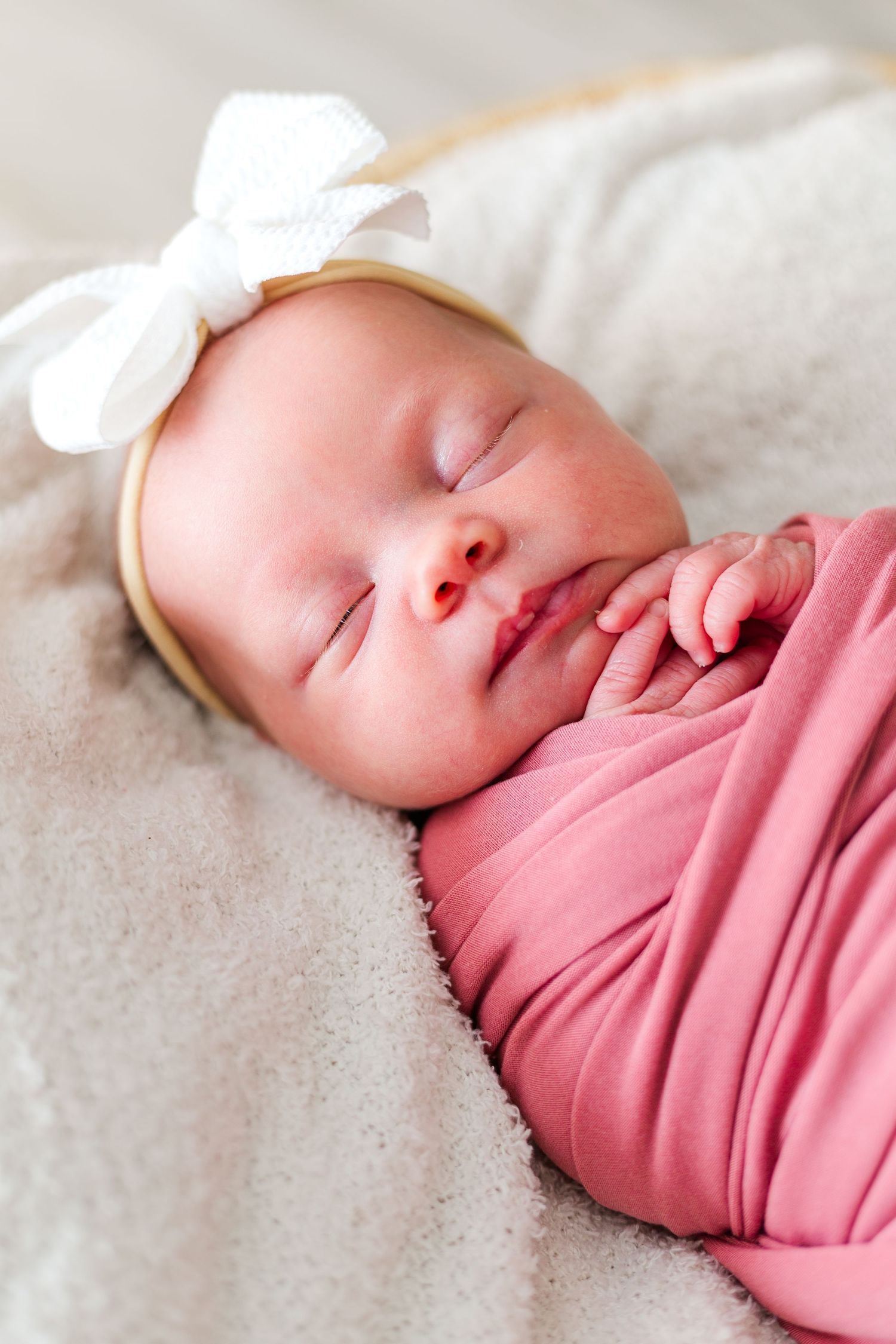 Sleeping newborn baby wrapped in pink swaddle wearing white bow headband resting on soft gray blanket.
