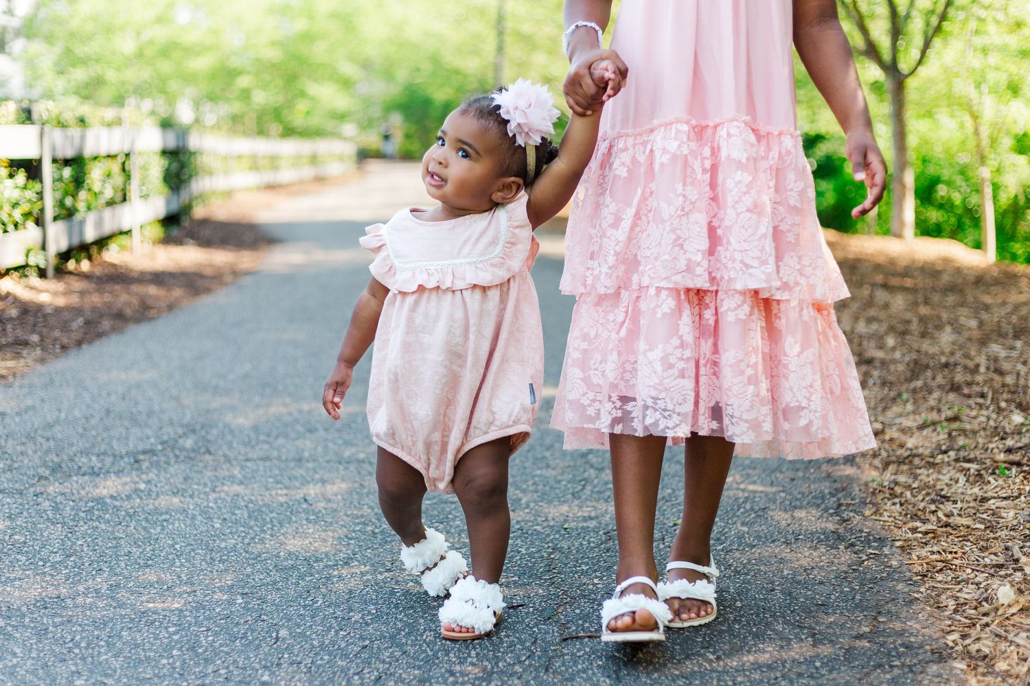 A toddler in pink romper holds hands with older girl in pink lace dress, walking on a garden path.